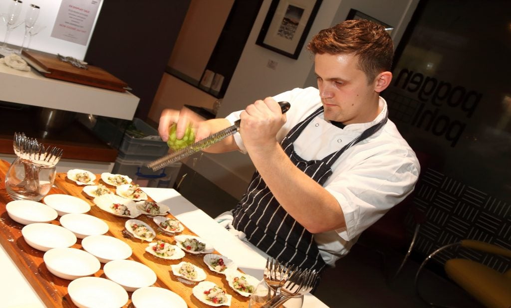 Chef Jon Watts preparing a dish in a modern kitchen. He wears a white chef’s coat with a black and white striped apron over it. He is grating a lime, directly over a platter of oysters that are neatly arranged on shells. The table is set with small white plates and utensils, ready for serving. The background includes some kitchen equipment, framed artwork on the walls, and a glass panel with partial text visible.