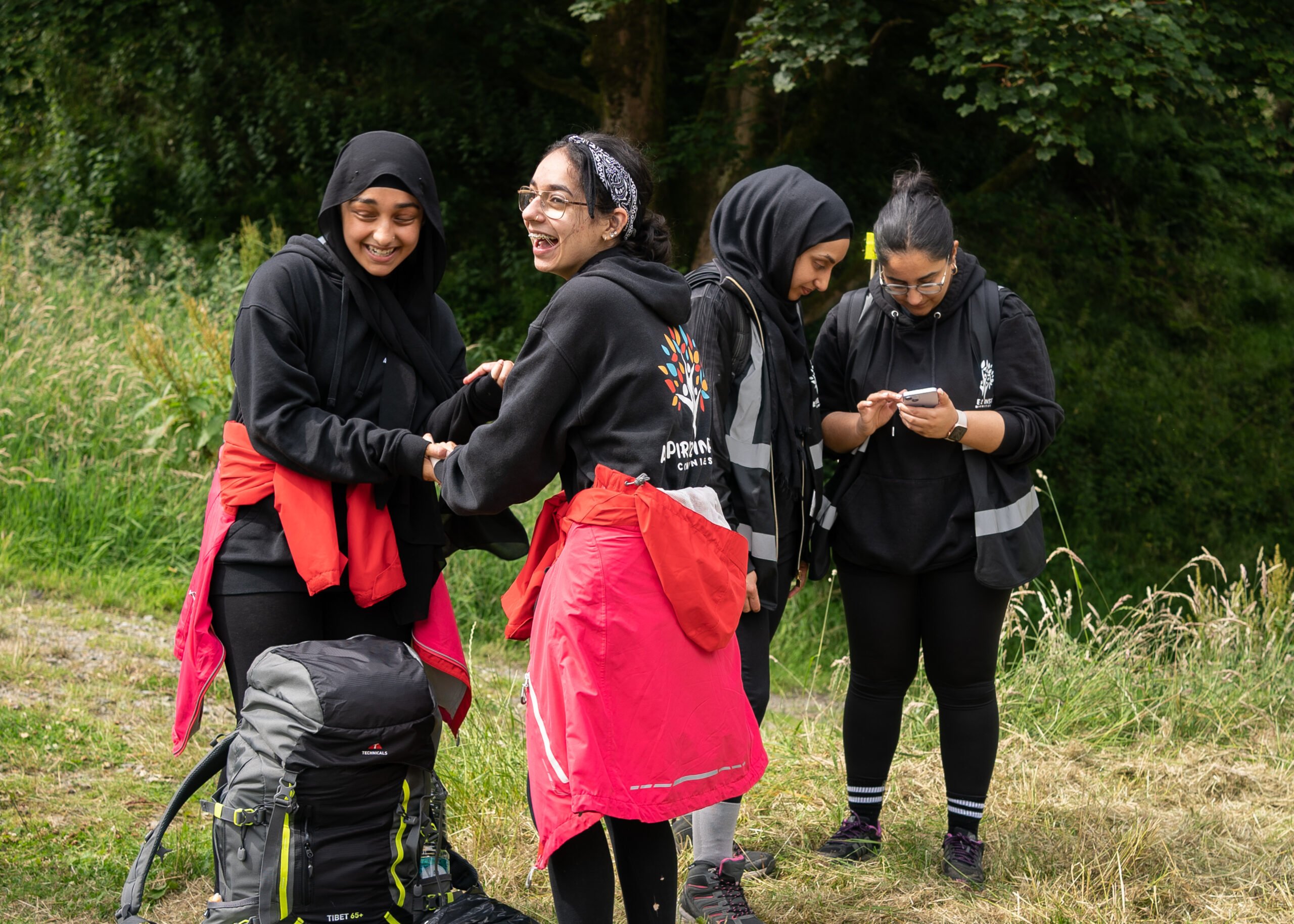 Four young people outside laughing together. They are all wearing rucksacks and hiking gear, 2 of them are looking at something on a mobile phone.