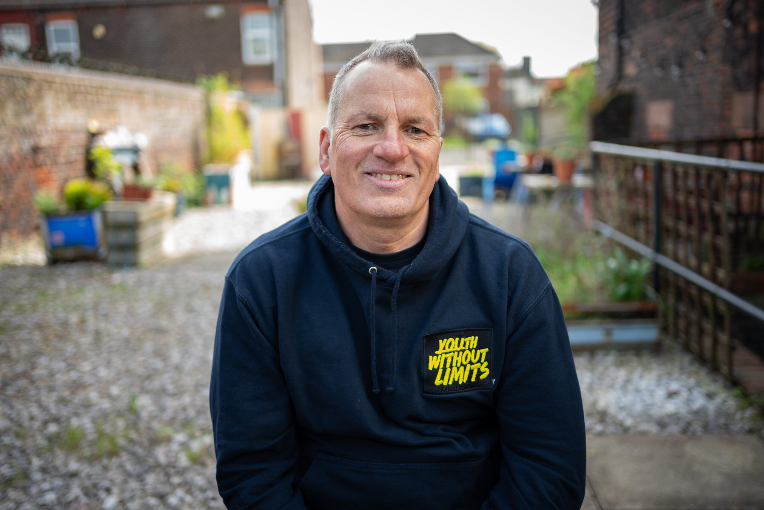 A man is smiling and staring straight at the camera. We can only see from his waist up and the background shows a pavement and some houses out of focus. He is wearing a navy "Youth Without Limits" DofE hoodie.