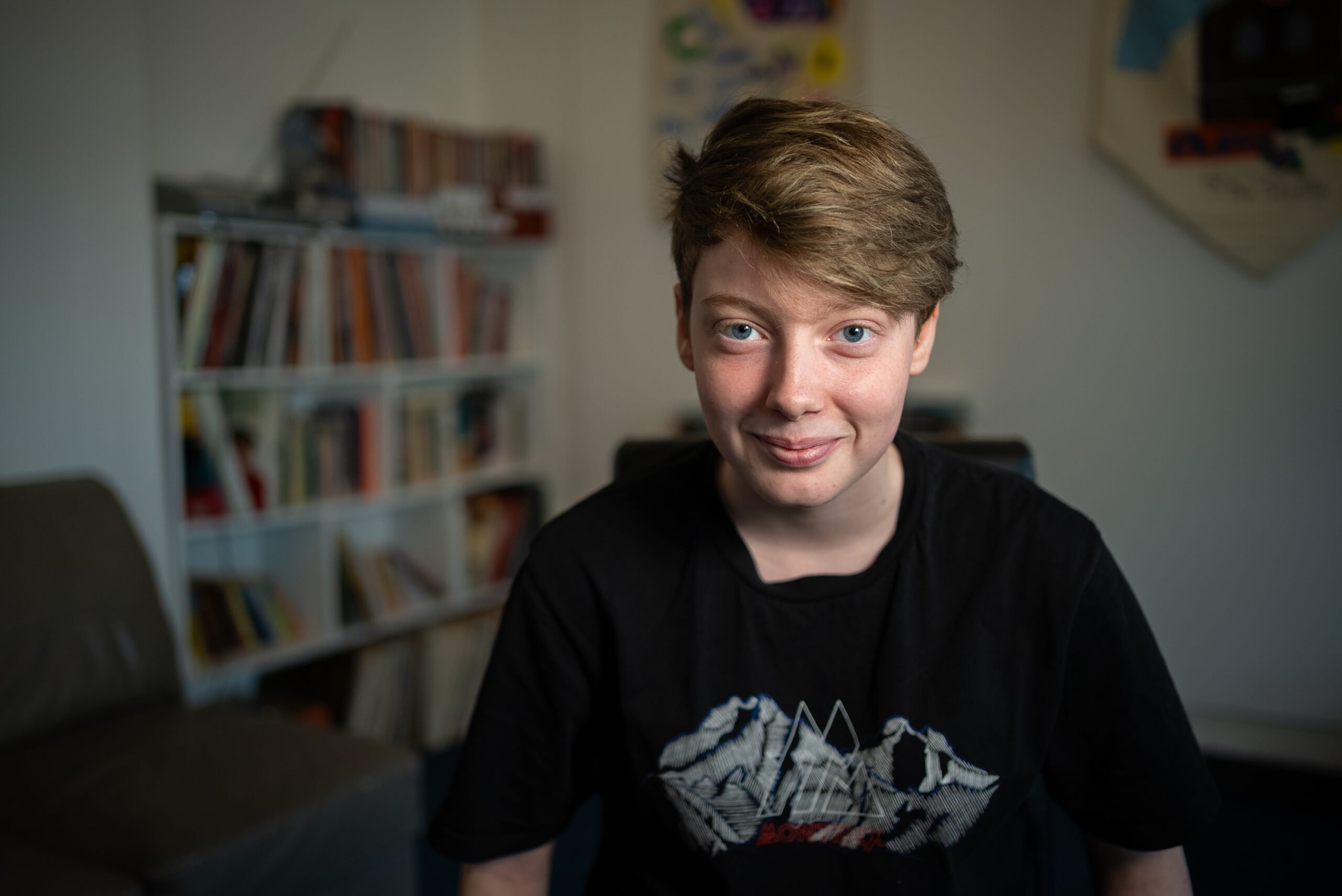 Ellesha smiles softly while sitting in a cosy room with bookshelves in the background. They are wearing a black t-shirt with a graphic design on the front. The lighting is warm, and the overall atmosphere of the room feels inviting and relaxed.