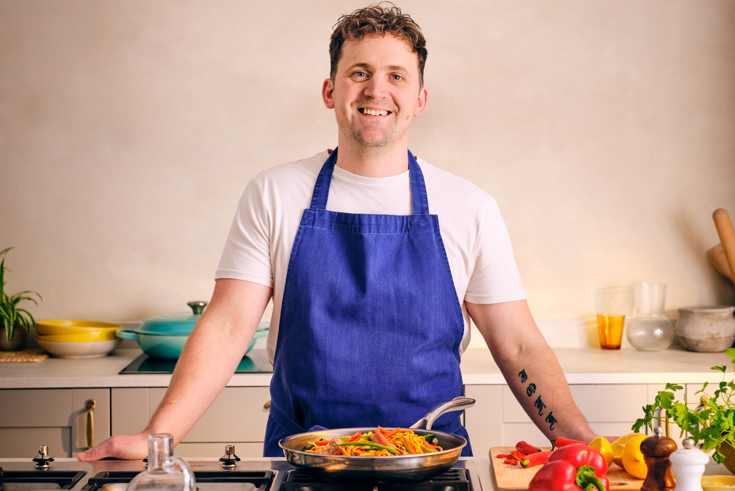 Jon Watts stands in a modern kitchen wearing a blue apron over a white t-shirt. He is positioned behind a stovetop, where a pan filled with colourful sautéed vegetables is cooking. On the countertop, there are various fresh ingredients including a red bell pepper, lemon, and herbs, along with a bottle of olive oil and some utensils. The background features a light-colored wall, minimalistic decor, and a few kitchen items like bowls and jars, adding to the bright and inviting atmosphere.