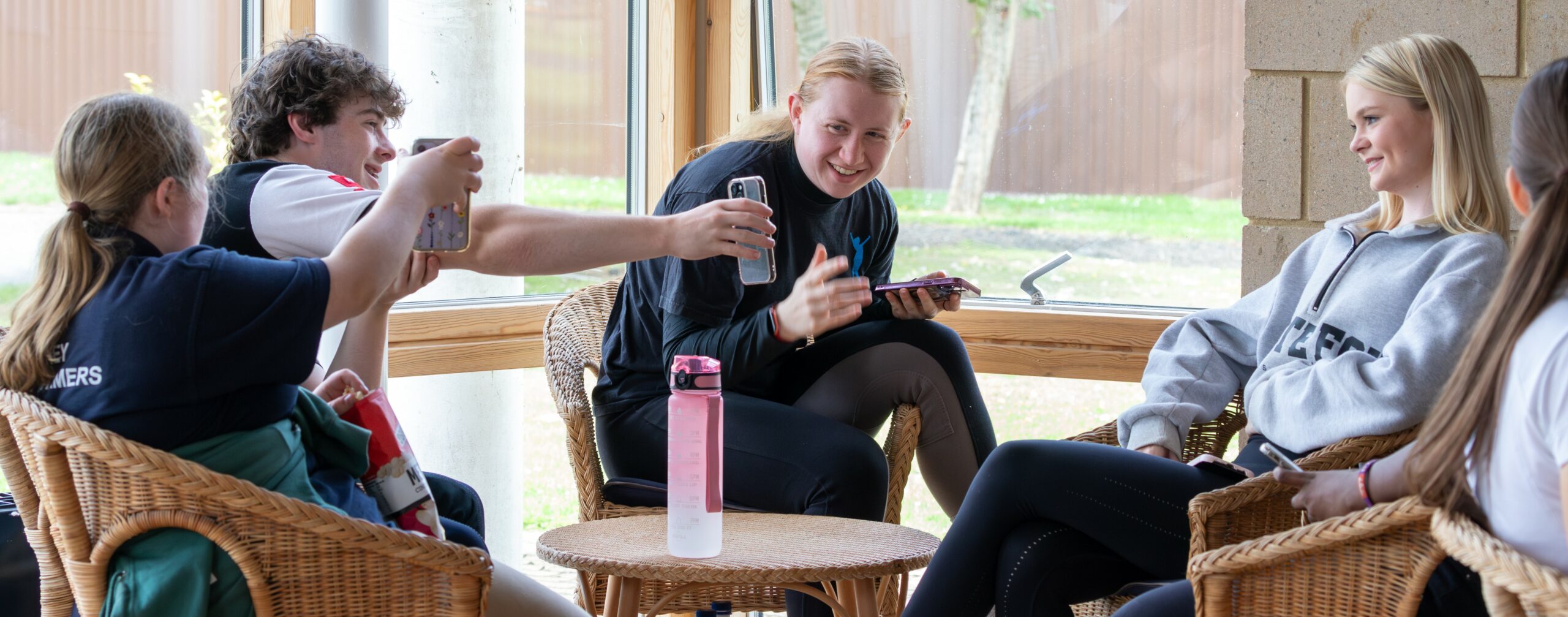 A group of young people sitting in a bright, airy space with large windows. Two individuals are engaged in conversation, while one person is taking a selfie. The atmosphere is relaxed and friendly, with wicker chairs and a small table in the center. Green trees are visible outside the windows, adding to the cheerful ambiance.