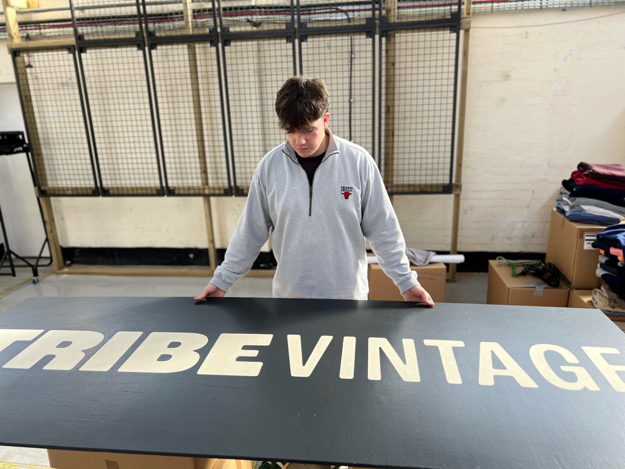 Freddie wearing a light grey sweatshirt with a small logo on the chest is standing indoors, looking down at a large black sign that reads 'TRIBE VINTAGE' in bold white letters. The setting appears to be a workspace, with cardboard boxes and folded clothing visible in the background."