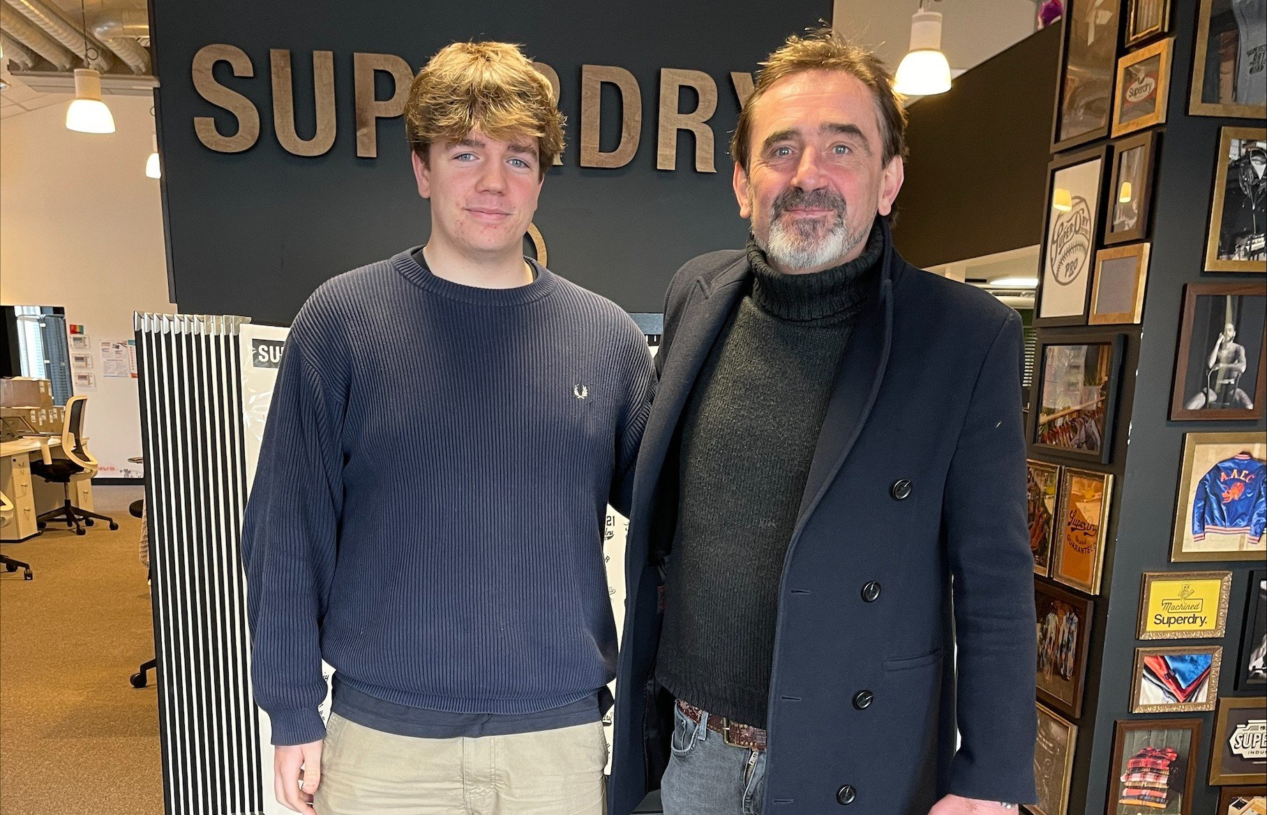 Freddie wearing a navy sweater, beige trousers, and green trainers stands next to Julian Dunkerton wearing a black coat, black turtleneck, skinny jeans, and white trainers. They are posing for a photo inside a modern office space with a 'Superdry' sign on the wall and framed artwork in the background.