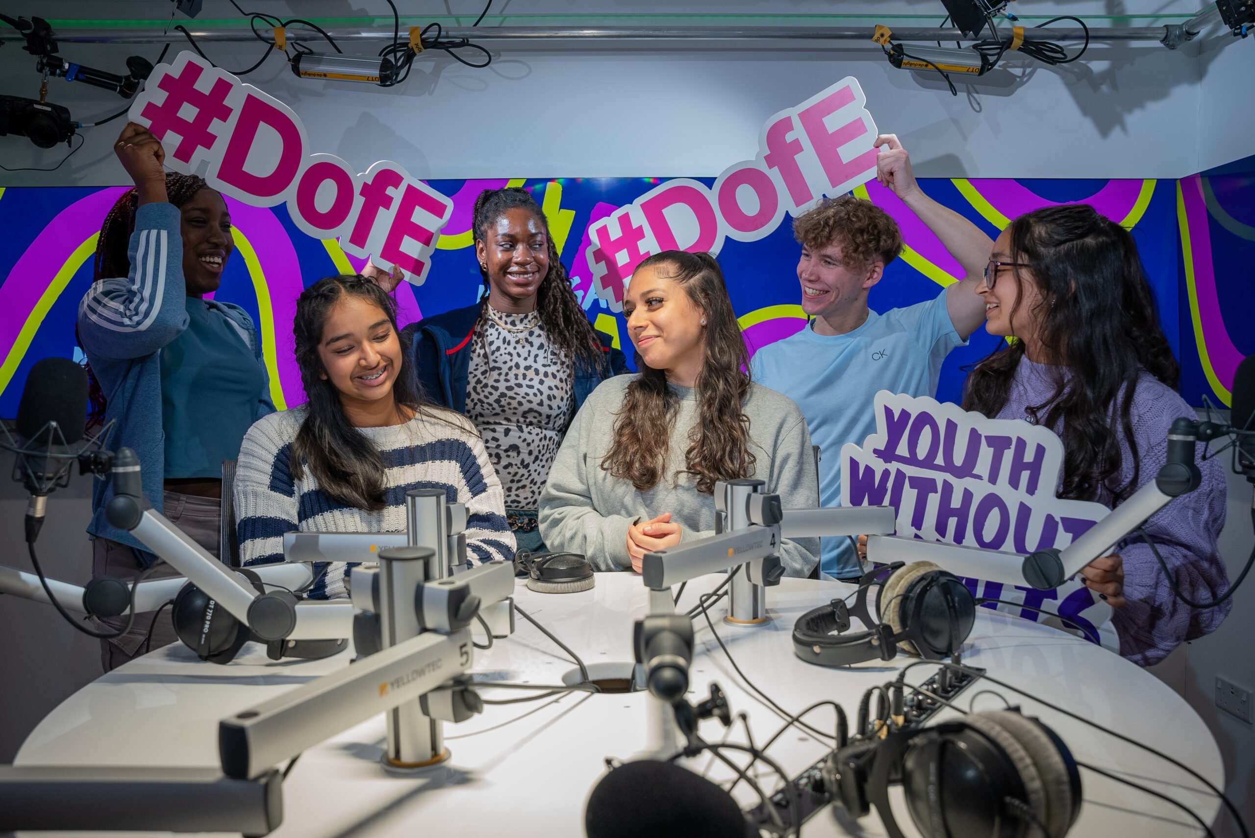 A diverse group of six young people is gathered in a podcast recording studio, smiling and holding up colorful signs that say "#DofE" and "Youth Without Limits." The group is surrounded by professional microphones, headphones, and equipment, with a vibrant, colorful backdrop behind them.