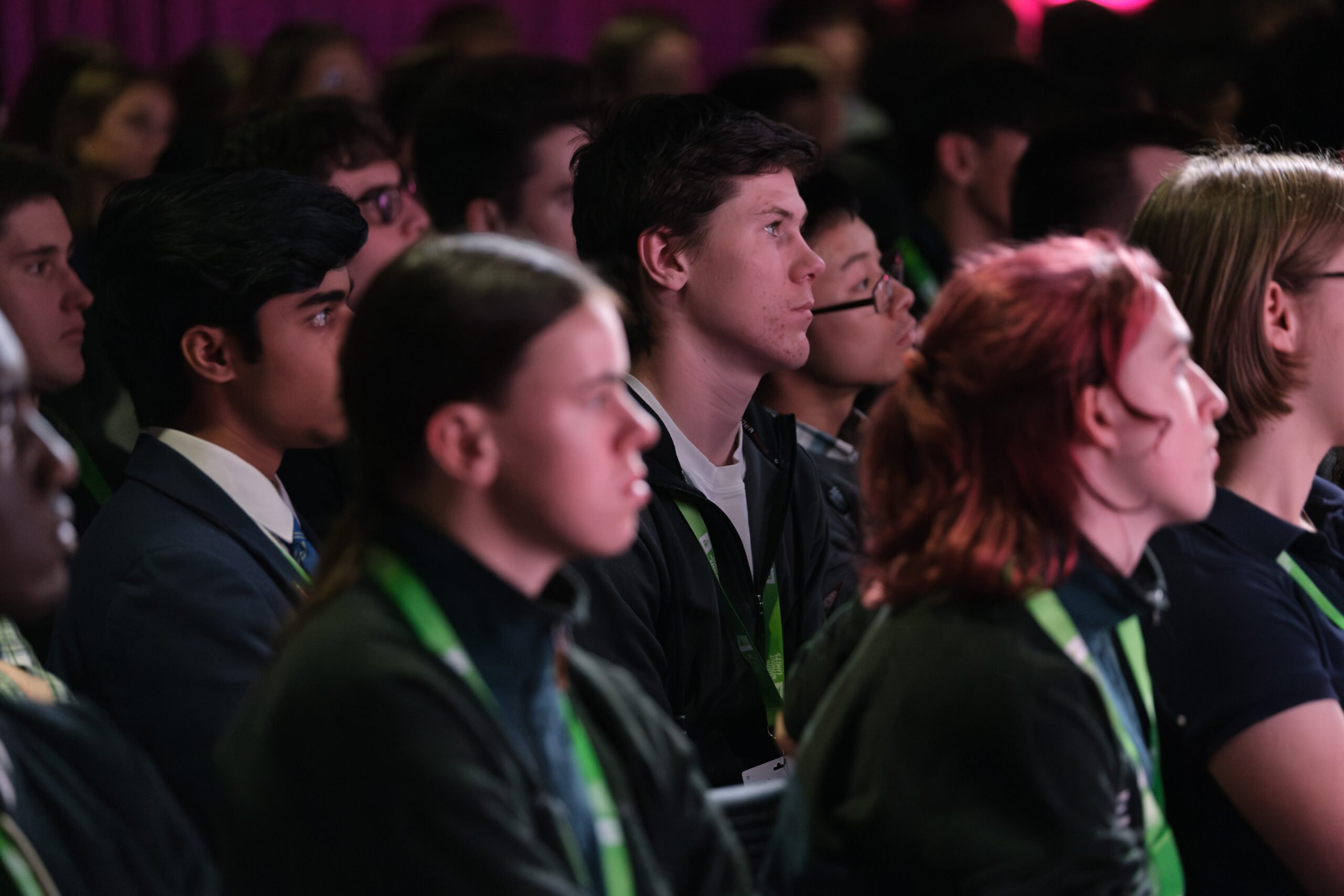 A diverse group of young people attentively listening during a presentation or event. They are seated in a semi-circle, with expressions of focus and engagement. Some individuals are wearing lanyards, and the background features soft lighting, creating a vibrant atmosphere.