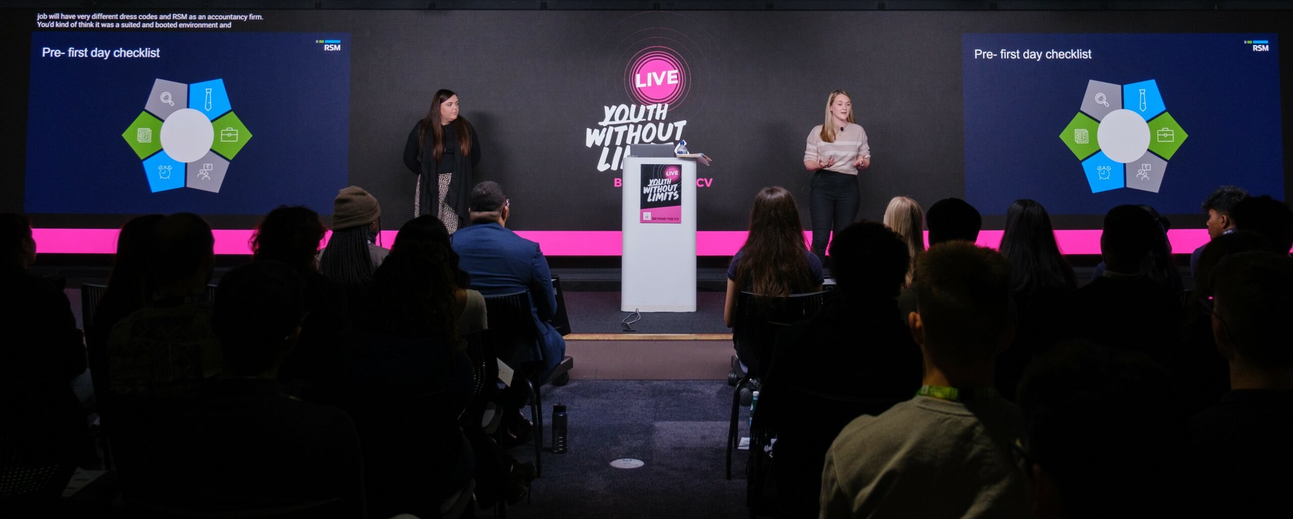 Two women stand on stage in front of a large screen displaying a "Pre-first day checklist." The setting is a modern conference room with an audience seated in front. The stage features a podium with a sign that reads "LIVE WITHOUT LIMITS." The atmosphere is professional and engaging.