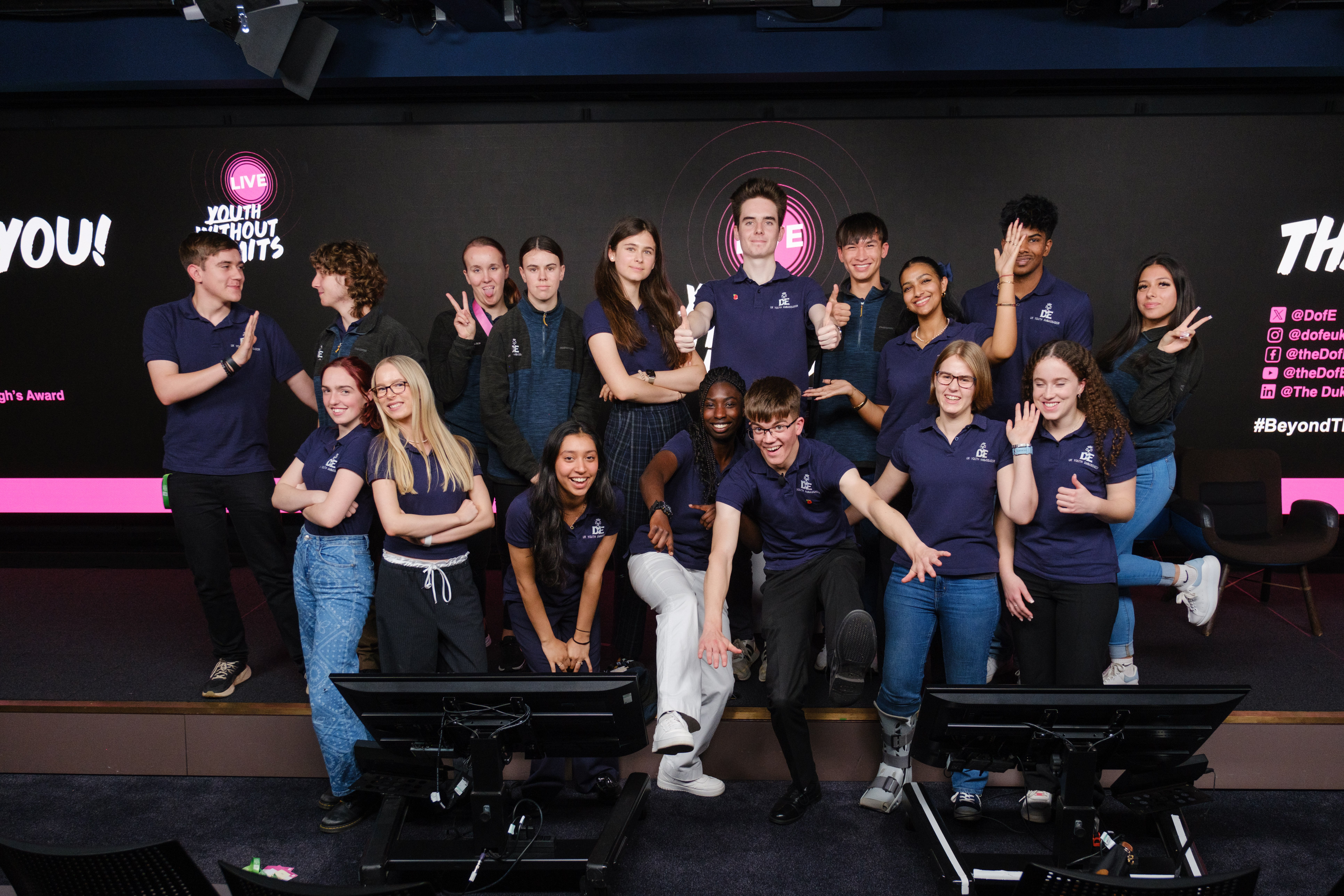 A diverse group of young people poses together in a modern event space, smiling and making playful gestures. They are wearing matching navy blue shirts and are arranged in a dynamic formation, with some individuals standing and others kneeling or sitting. The background features a large screen with the text "Thank You!" and social media handles, creating a vibrant and energetic atmosphere.