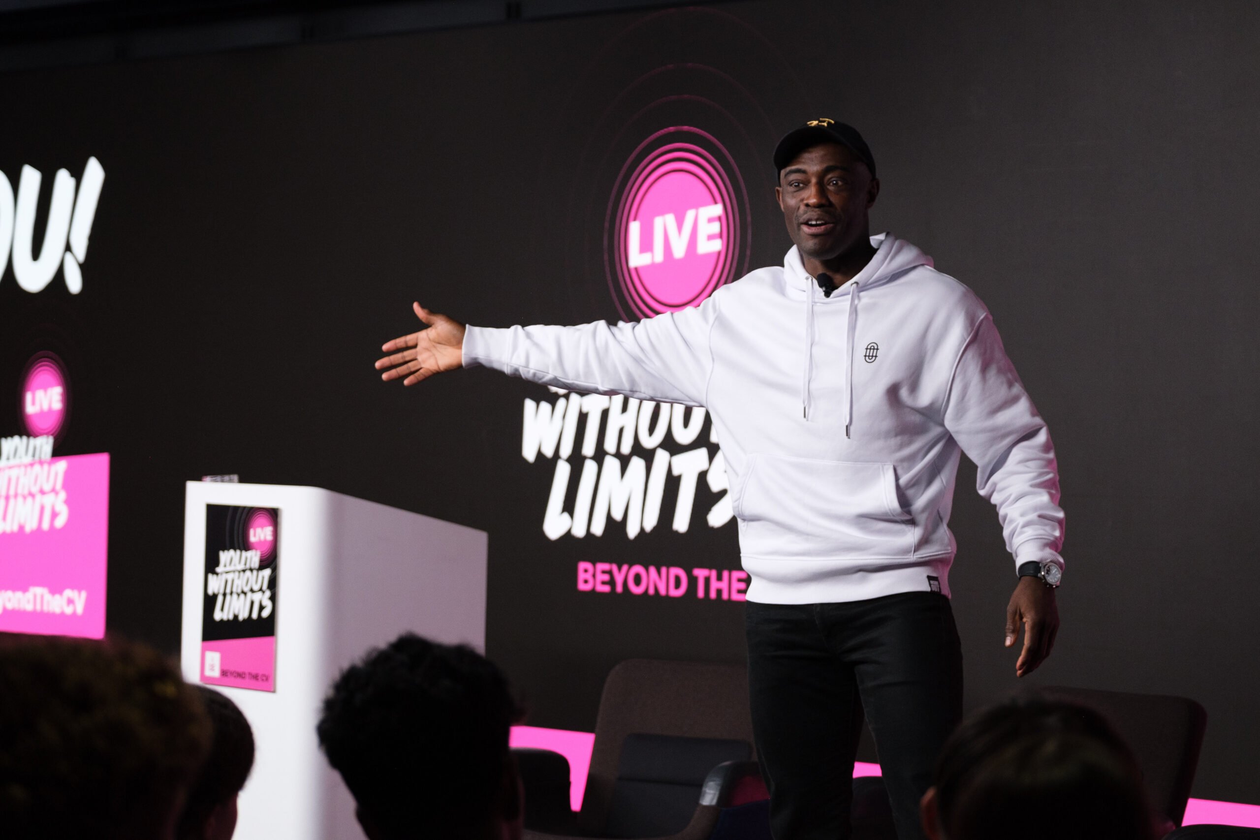 Tim Campbell MBE delivers an inspiring speech on stage at the Youth Without Limits Live - Beyond the CV event. He is wearing a white hoodie and a black cap, gesturing with one arm extended as he addresses the audience. A large screen behind him displays the event branding, with bold text that reads 'Youth Without Limits' and 'Beyond the CV.' Audience members are visible in the foreground.