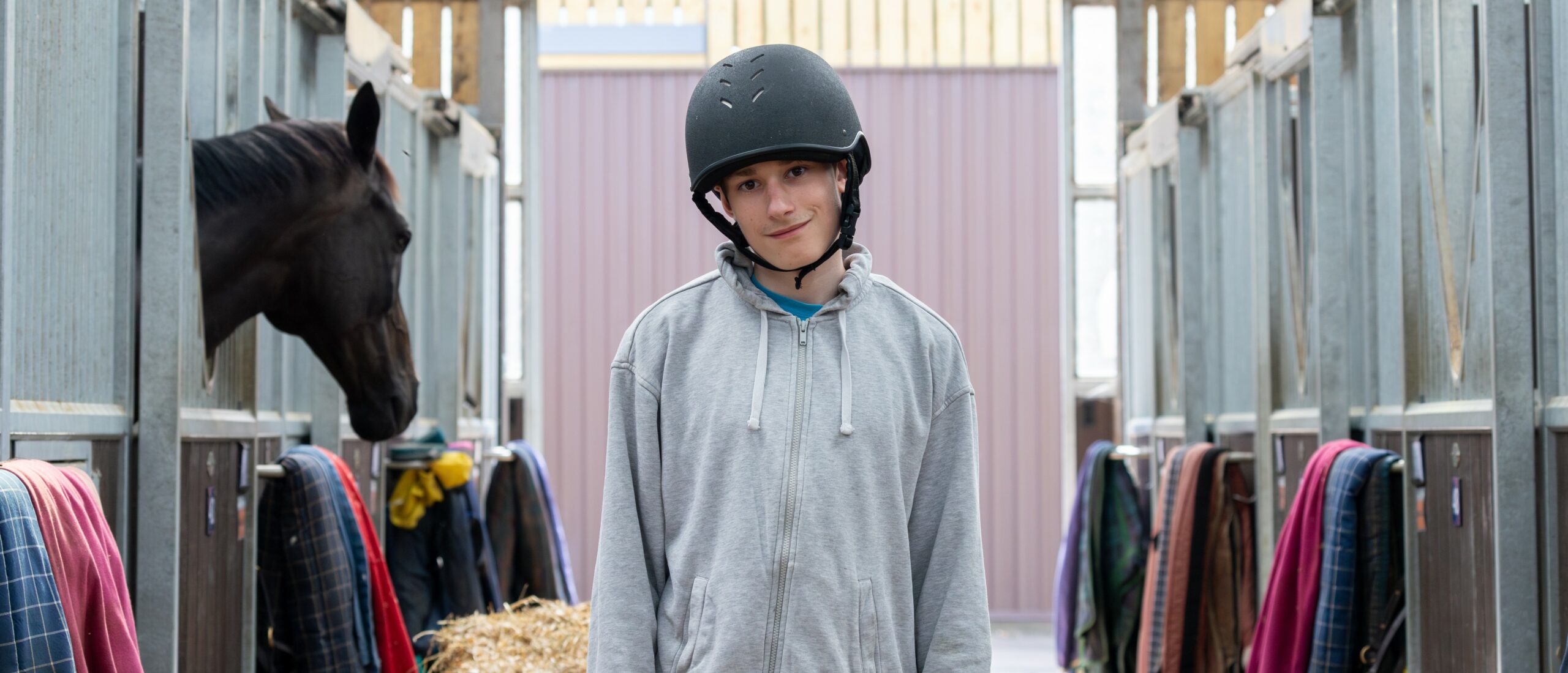 Ethan-Thomas wearing a helmet stands in a stable, surrounded by horse stalls. One horse's head is visible in the stall to the left. The stalls are lined with colorful blankets and hay, and the stable has wooden beams and a bright background. The person has a relaxed expression and is dressed in casual clothing.