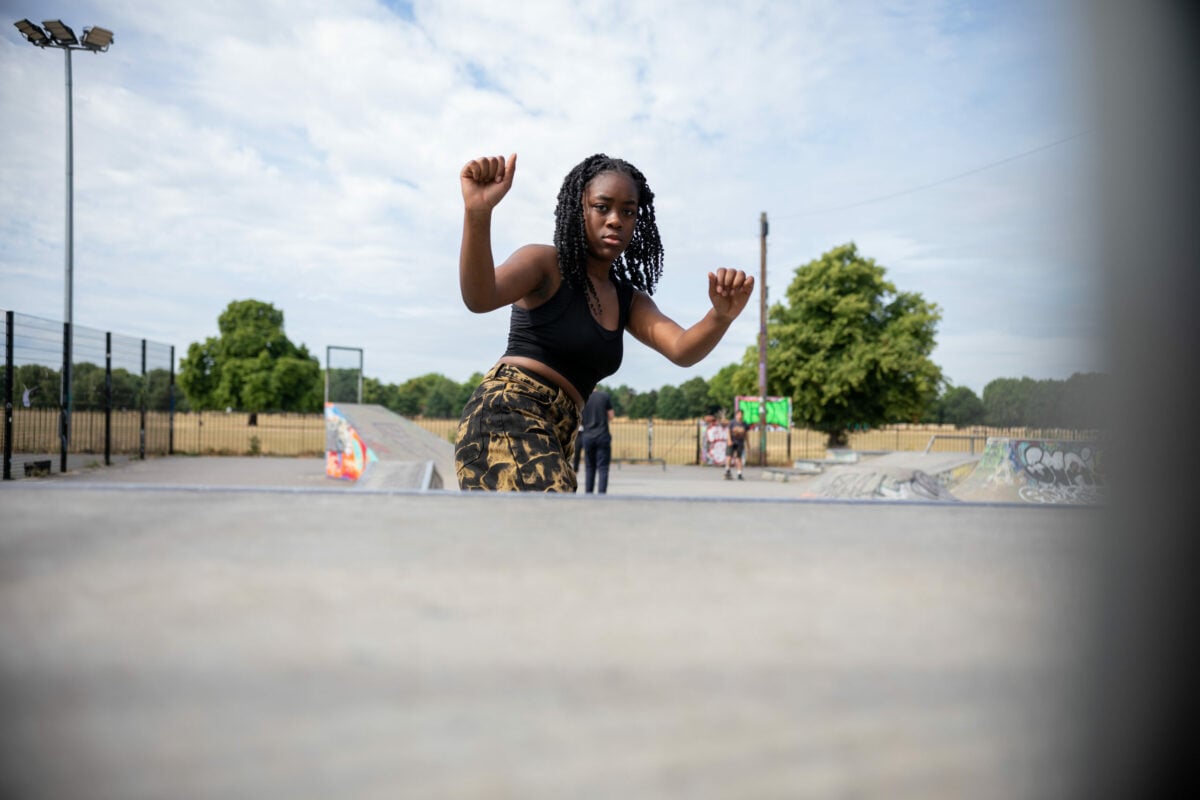 A young woman with braided hair wearing a black crop top and patterned trousers poses confidently in a skate park. The background features ramps, graffiti, and green trees under a partly cloudy sky.