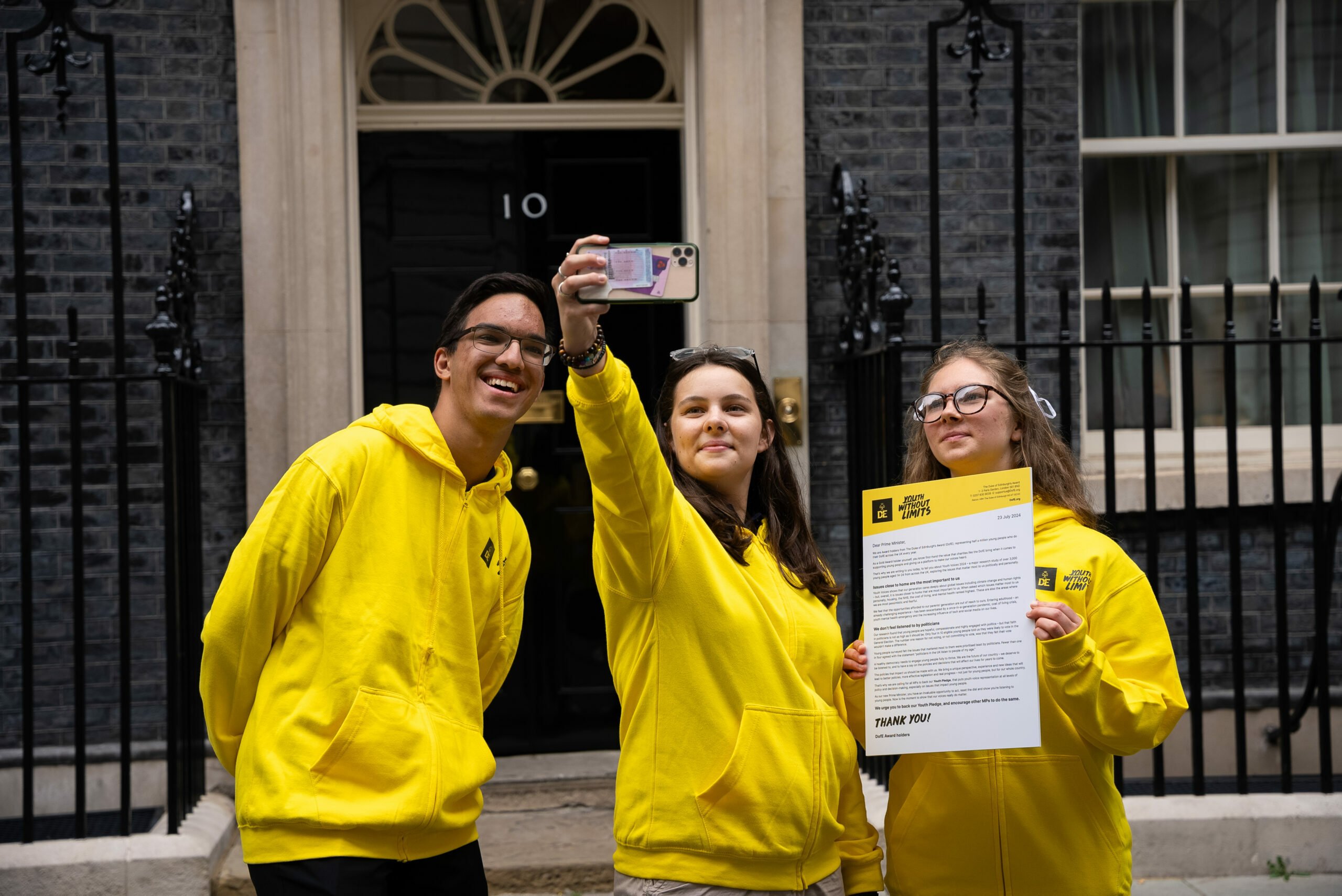 Three young people wearing yellow hoodies are standing in front of the iconic black door of 10 Downing Street. One of them is taking a selfie with a smartphone, while another is holding a document with the heading 