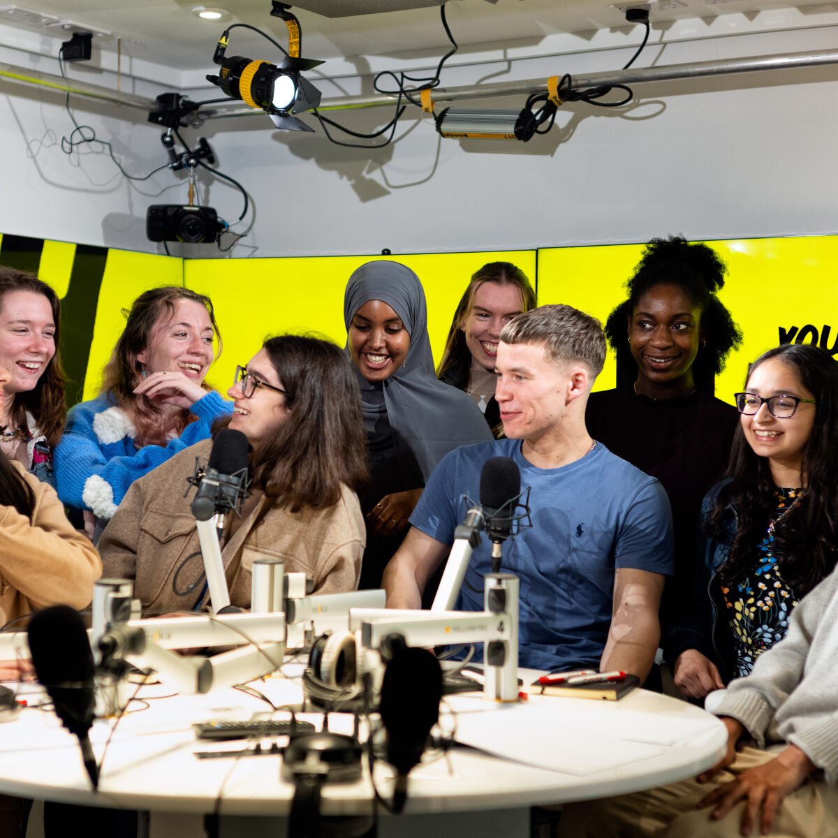 A group of young people sat around a table with microphones and podcast recording equipment. They are all smiling and laughing together, in the background we can see studio lights and a yellow backdrop on a large screen.