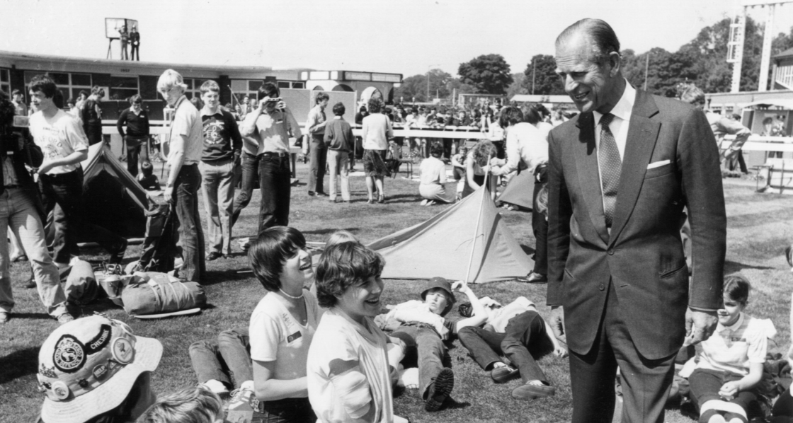 A large group of people, including children and adults, are gathered outdoors on a sunny day. In the foreground, an older gentleman in a suit is smiling and interacting with a group of children who are sitting and lying on the grass, laughing and looking up at him. The background shows a busy scene with many people, some setting up tents and others engaged in various activities, giving the impression of a lively event or festival.