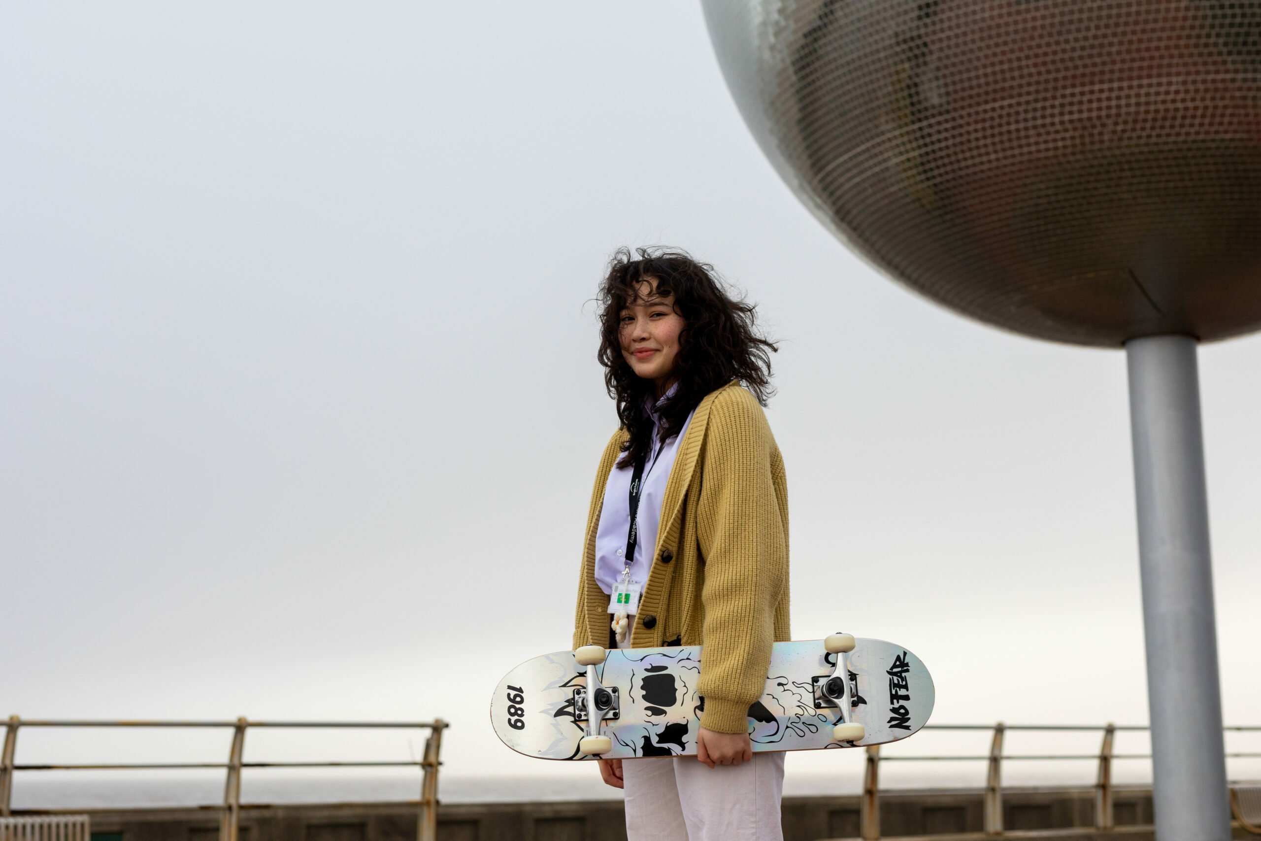 A young woman stands outdoors holding a skateboard. She is wearing a yellow cardigan over a white shirt and light-colored pants. She has curly hair and is smiling at the camera. Behind her is a railing, a cloudy sky, and a large metallic sphere sculpture.