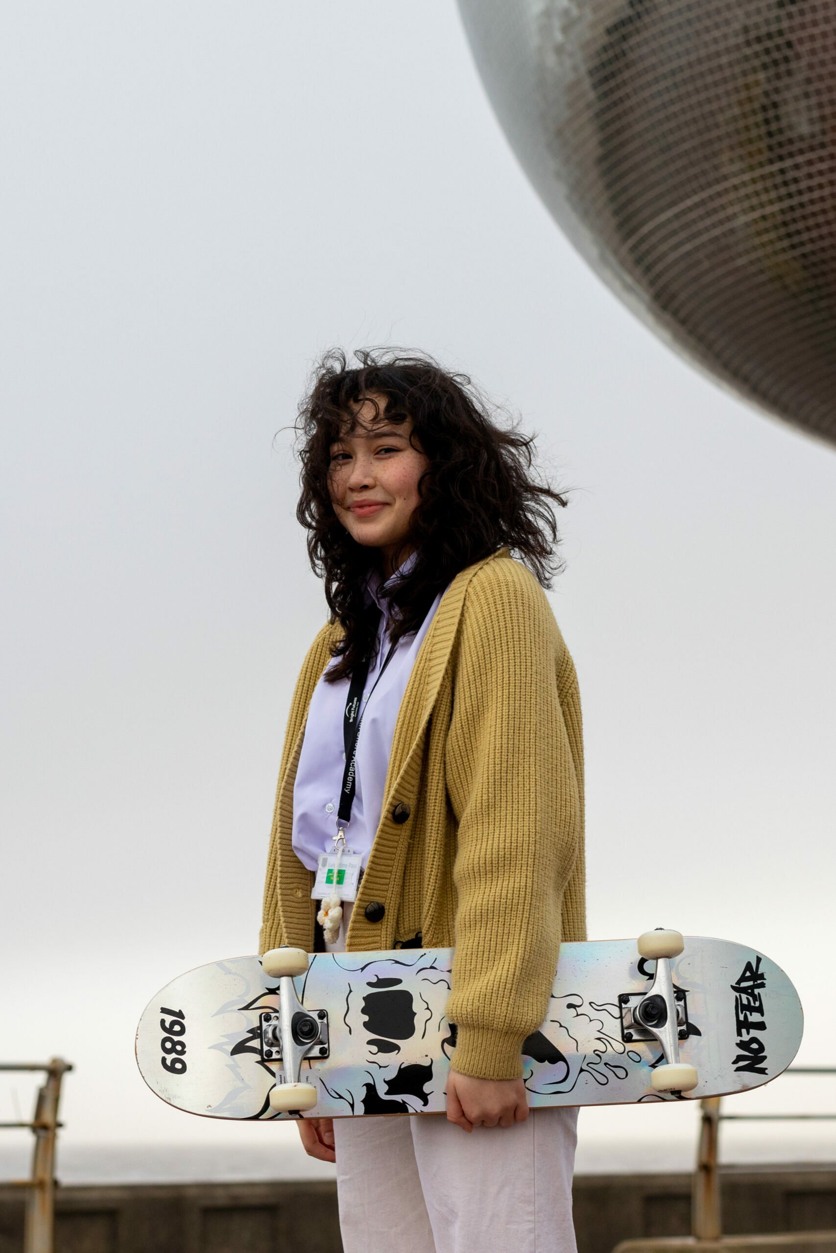 A young woman stands outdoors holding a skateboard. She is wearing a yellow cardigan over a white shirt and light-colored pants. She has curly hair and is smiling at the camera. Behind her is a railing, a cloudy sky, and a large metallic sphere sculpture.