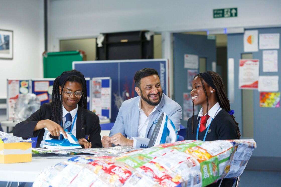 A man in a light blue blazer is seated at a table with two young women in school uniforms. They are all smiling and appear to be engaged in a friendly conversation. One of the young women is holding an iron and working on a craft project with colorful materials spread out on the table. The background shows a classroom environment with posters and displays on the walls. The overall atmosphere is cheerful and collaborative.