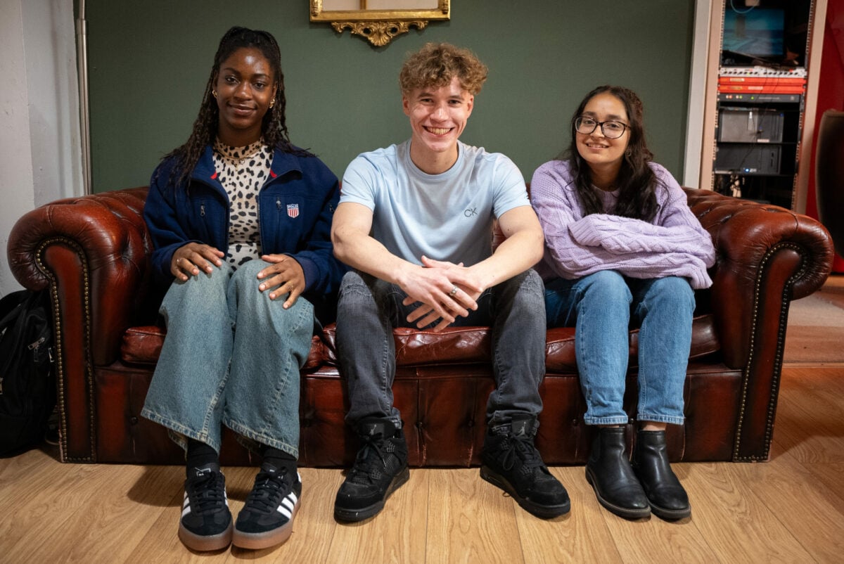 Three young people are sitting on a brown leather couch. From left to right: the first person is a woman with long braided hair, wearing a navy jacket, a leopard-print top, wide-leg jeans, and black Adidas sneakers; the middle person is a man with curly blonde hair, wearing a light blue T-shirt, dark jeans, and black sneakers, smiling with his hands clasped in front; the third person is a woman with long dark hair and glasses, wearing a light purple sweater, blue jeans, and black ankle boots, with her arms folded on her lap. The background features a green wall with a decorative golden frame