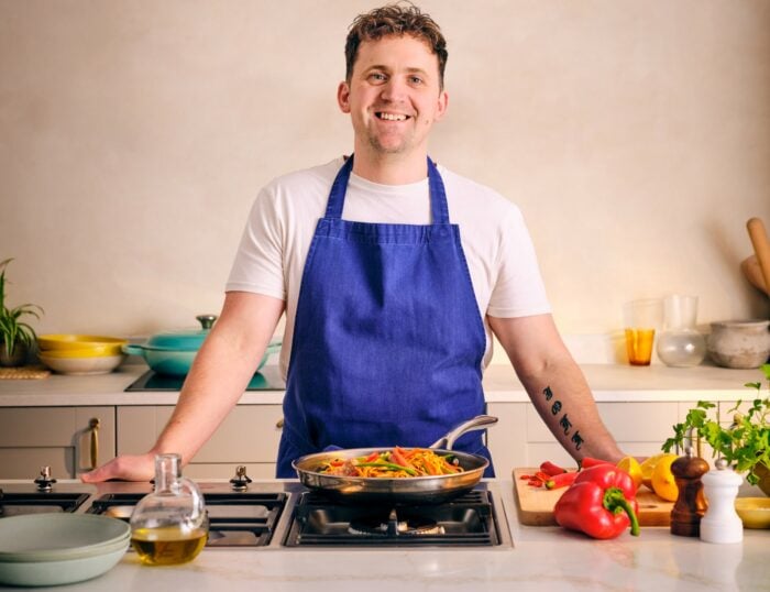 Jon Watts stands in a modern kitchen wearing a blue apron over a white t-shirt. He is positioned behind a stovetop, where a pan filled with colourful sautéed vegetables is cooking. On the countertop, there are various fresh ingredients including a red bell pepper, lemon, and herbs, along with a bottle of olive oil and some utensils. The background features a light-colored wall, minimalistic decor, and a few kitchen items like bowls and jars, adding to the bright and inviting atmosphere.