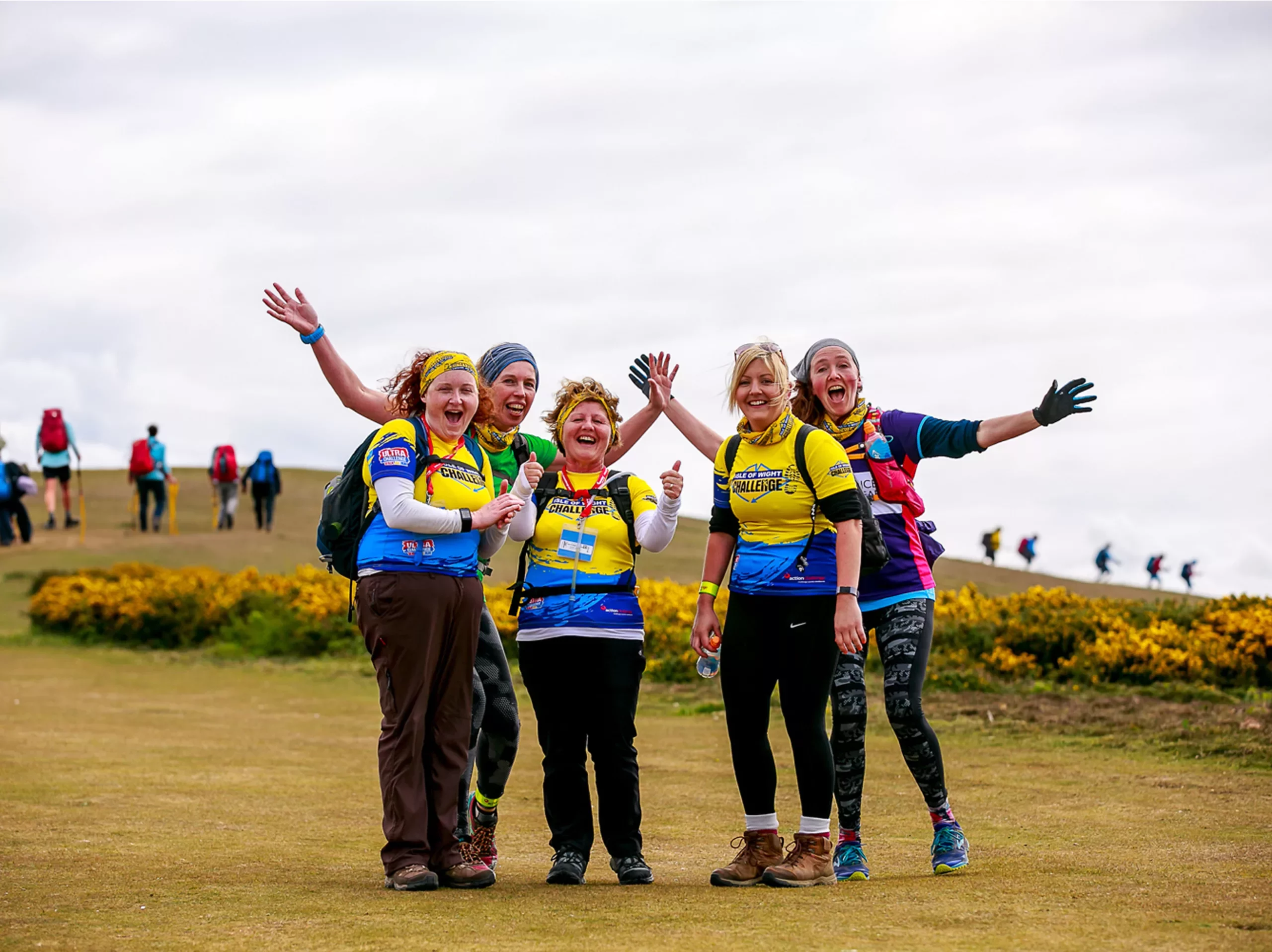 A group of five women standing outdoors on a grassy trail during what appears to be an organized challenge event. They are all wearing matching yellow and blue jerseys, with the words 