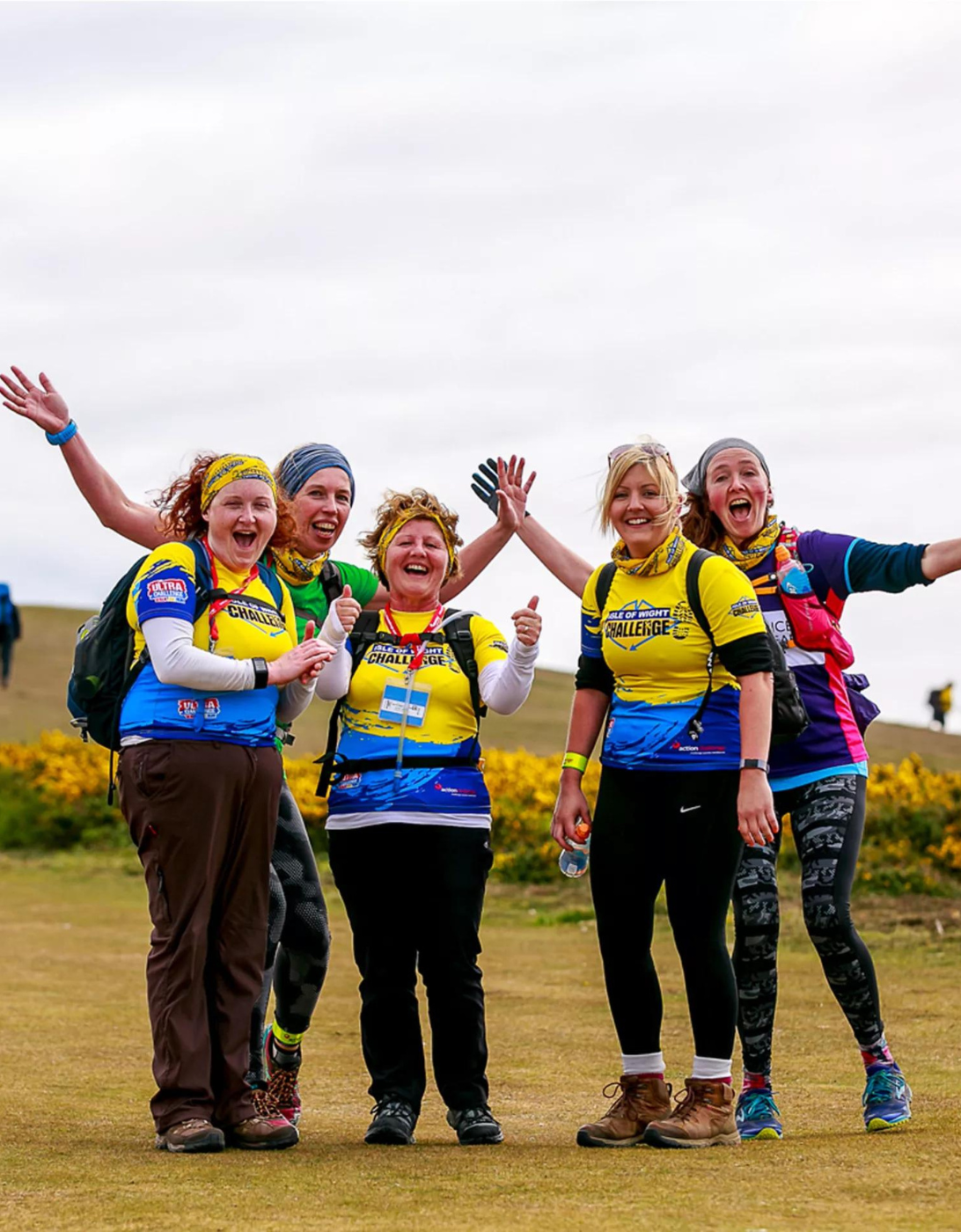 A group of five women standing outdoors on a grassy trail during what appears to be an organized challenge event. They are all wearing matching yellow and blue jerseys, with the words 