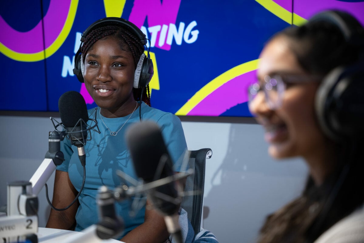 Podcast host, Elsie, is seated at a podcast recording table, wearing headphones and speaking into a microphone. She is smiling and appears to be engaged in the conversation. In the foreground, another person is slightly blurred but also smiling and wearing headphones. The background features a colorful display with abstract shapes and partial text that reads 