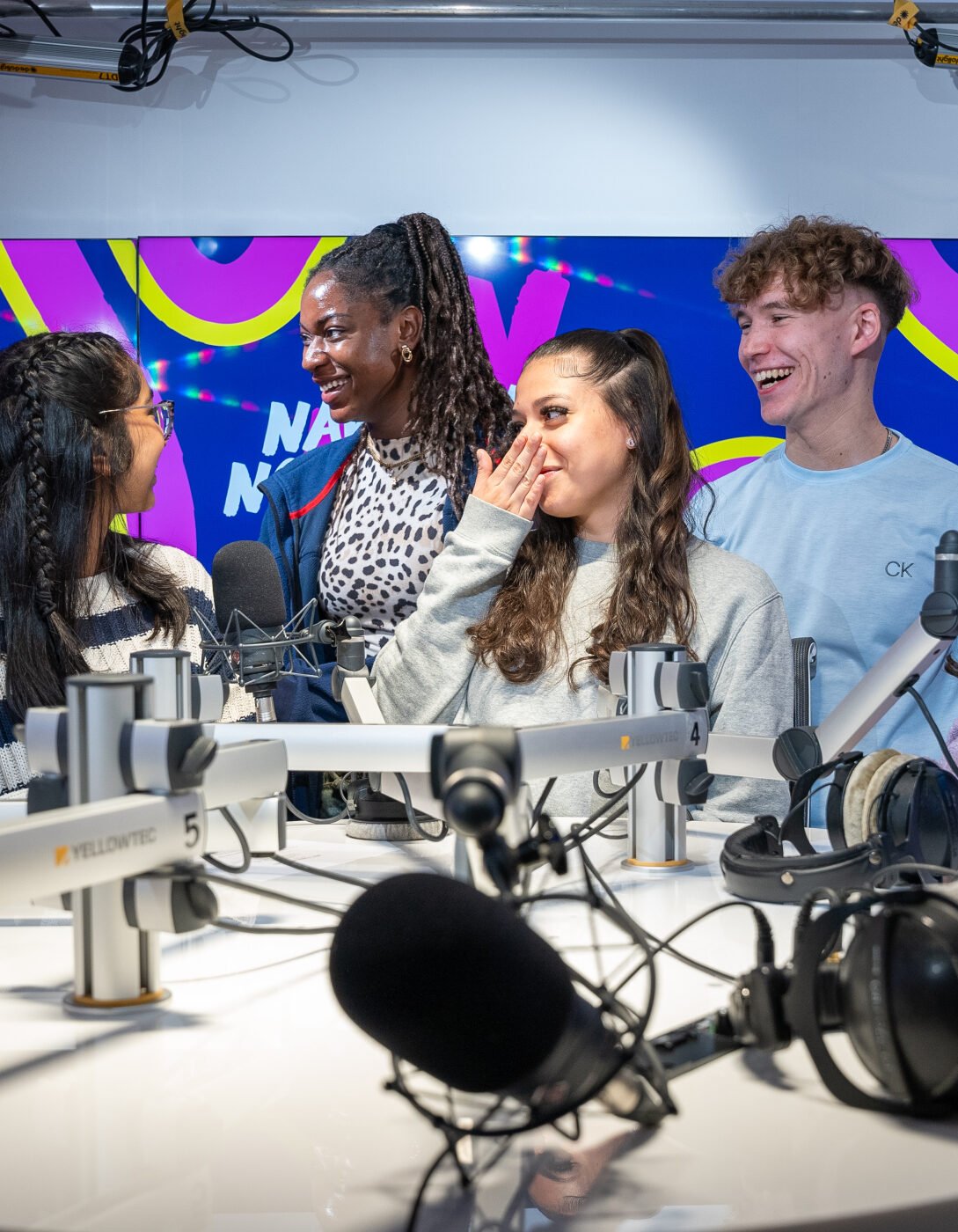 Six young people are gathered around a table with microphones in a recording studio. They are all smiling, laughing, and engaging with each other. The studio has bright lighting, and a colorful background display with the words 