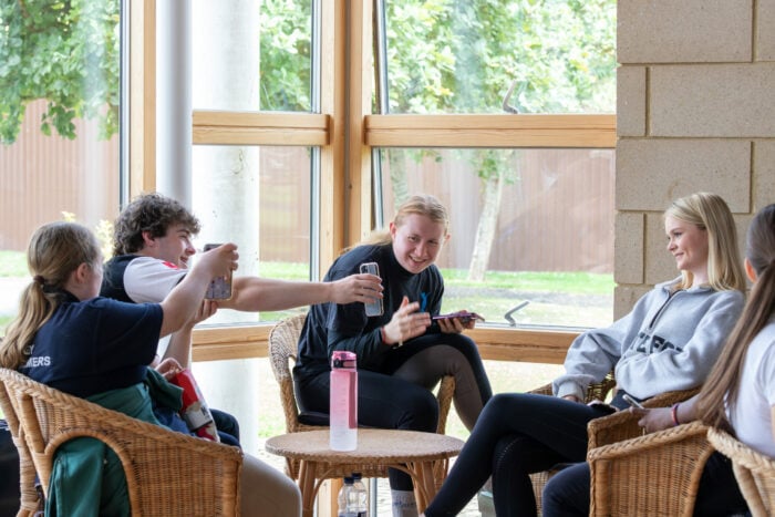 A group of young people sitting in a bright, airy space with large windows. Two individuals are engaged in conversation, while one person is taking a selfie. The atmosphere is relaxed and friendly, with wicker chairs and a small table in the center. Green trees are visible outside the windows, adding to the cheerful ambiance.