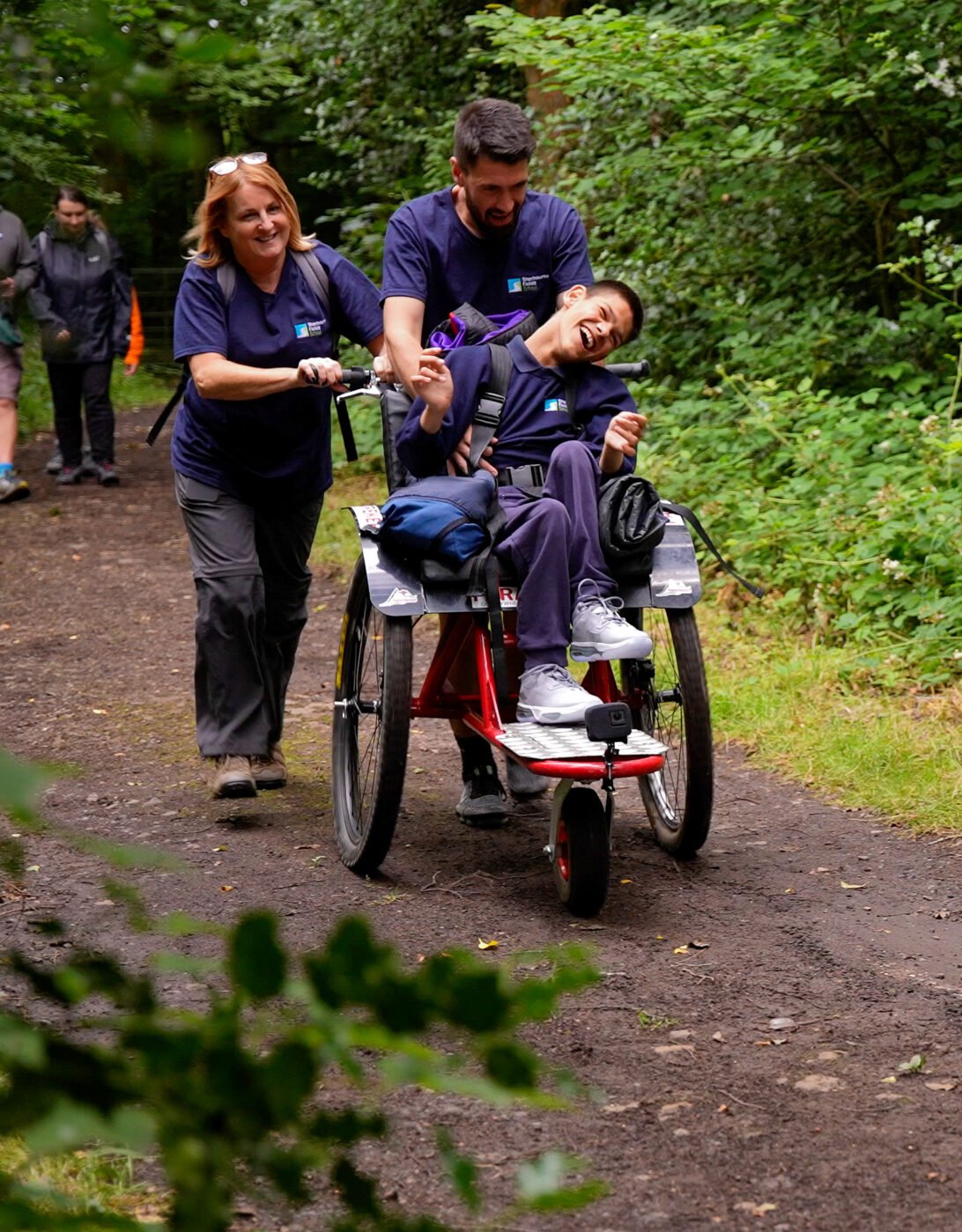 Two DofE Leaders assist Taylor in his wheelchair on a forest path. The woman is pushing the wheelchair while the man is smiling and engaging with Taylor. Lush greenery surrounds the path, and other individuals can be seen walking in the background.