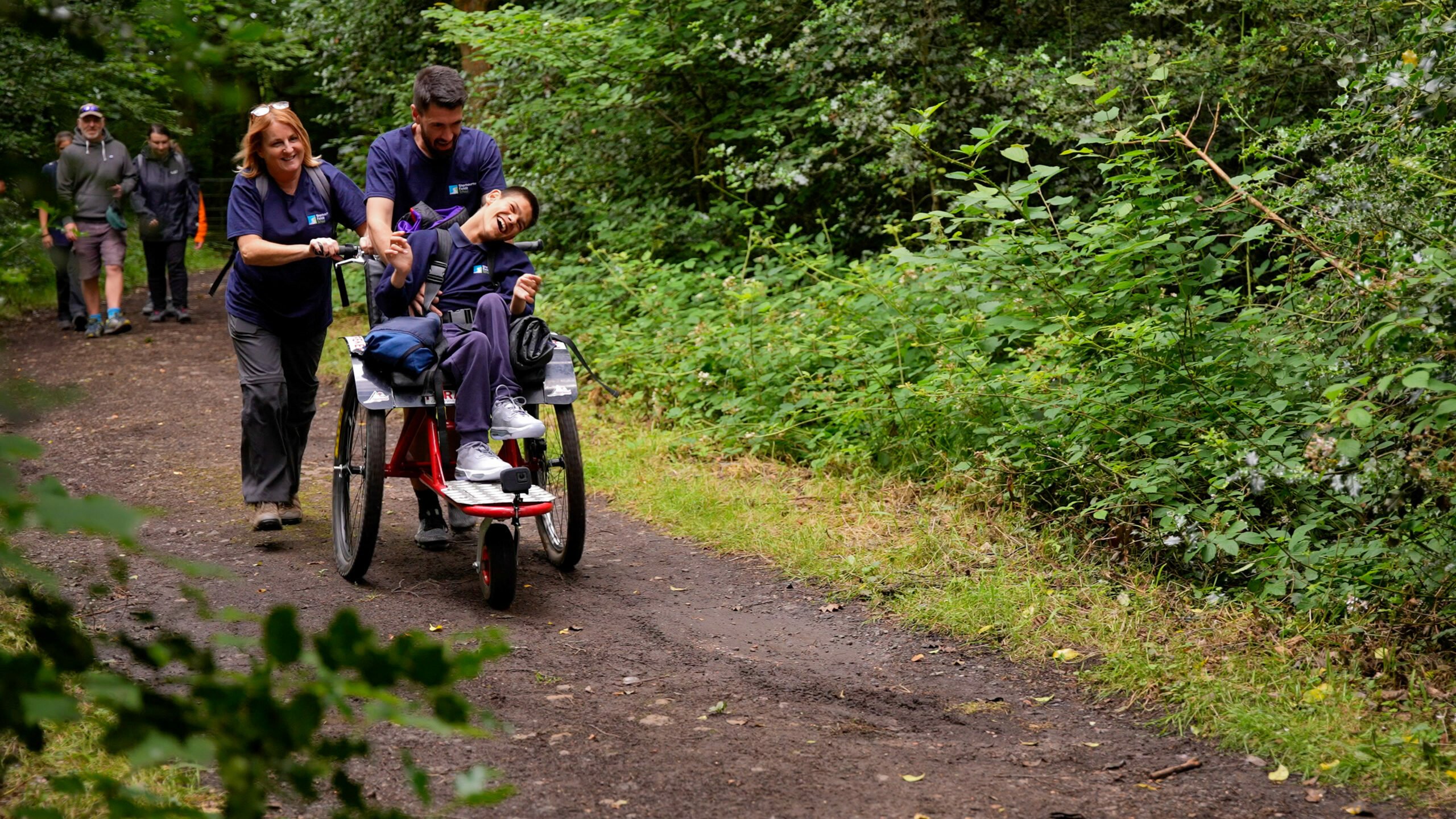 Two DofE Leaders assist Taylor in his wheelchair on a forest path. The woman is pushing the wheelchair while the man is smiling and engaging with Taylor. Lush greenery surrounds the path, and other individuals can be seen walking in the background.