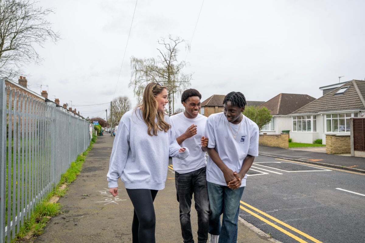 A group of three teenagers is walking down a residential street on a cloudy day. The two boys and one girl are dressed casually in light-colored sweatshirts and T-shirts. They appear to be enjoying each other's company, laughing and talking. The scene is set along a suburban neighborhood with houses, a fenced path on one side, and bare trees in the background,