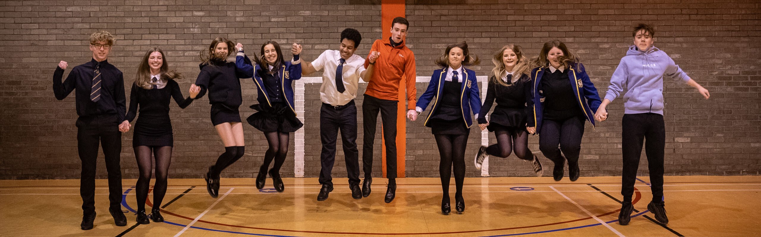 A group of nine young people are mid-jump in a school gymnasium, holding hands and smiling. The students are dressed in various outfits: some are in school uniforms with blue blazers, white shirts, and black skirts or trousers, while others are in more casual attire like hoodies and sweaters. They are all facing forward with an energetic and joyful demeanor. The gymnasium has a wooden floor with sports markings and a brick wall in the background, featuring a vertical orange stripe.