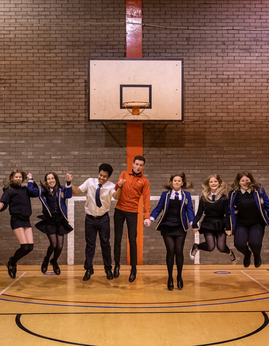 A group of nine young people are mid-jump in a school gymnasium, holding hands and smiling. The students are dressed in various outfits: some are in school uniforms with blue blazers, white shirts, and black skirts or trousers, while others are in more casual attire like hoodies and sweaters. They are all facing forward with an energetic and joyful demeanor. The gymnasium has a wooden floor with sports markings and a brick wall in the background, featuring a vertical orange stripe.