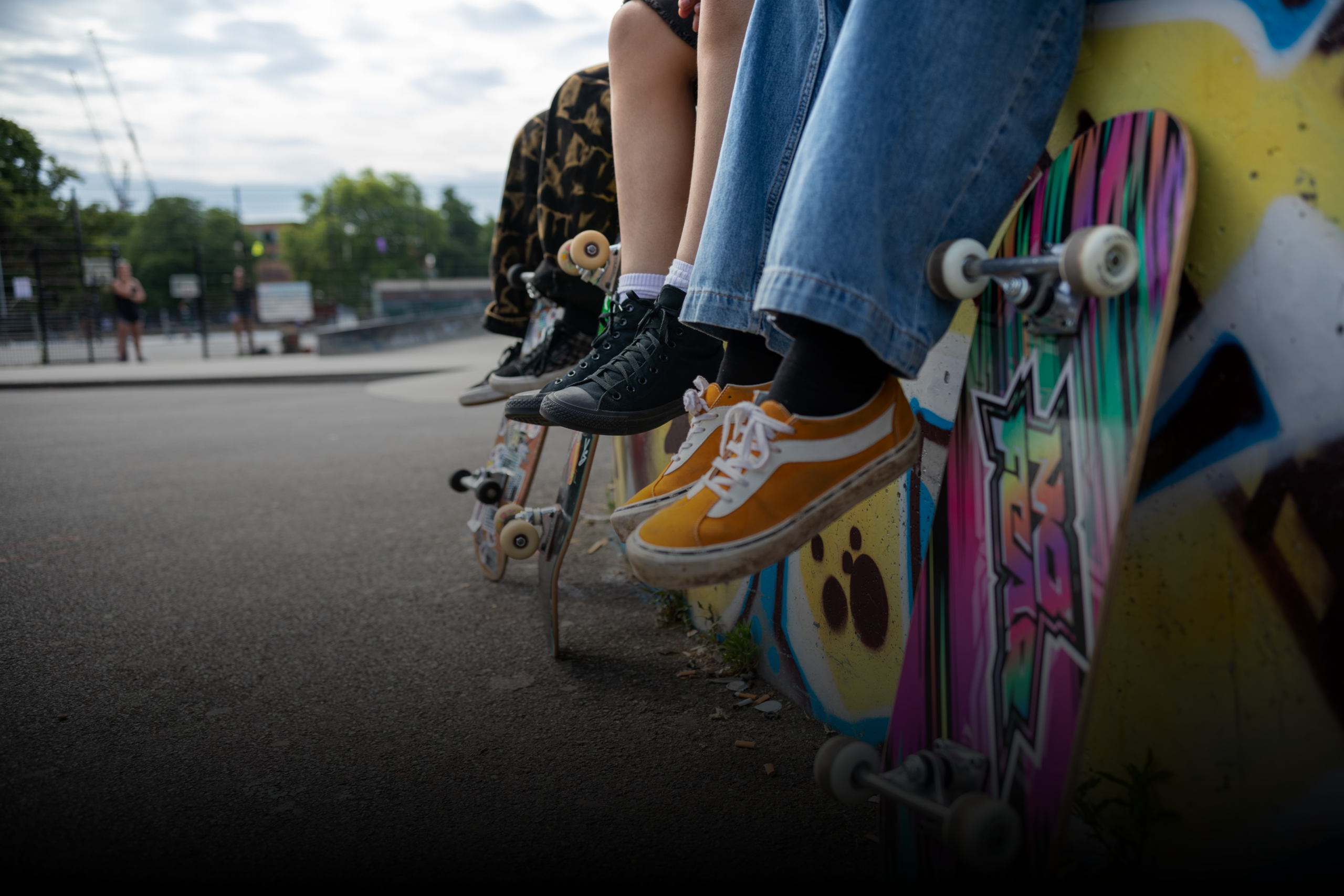 A close-up of a skateboard leaning against a wall with colorful graffiti. A person wearing orange and white sneakers and blue wide-legged jeans is sitting on the edge of the wall, with their feet dangling above the pavement. The skateboard has vibrant, multicolored stripes and visible white wheels. Other individuals and skateboards are slightly blurred in the background.