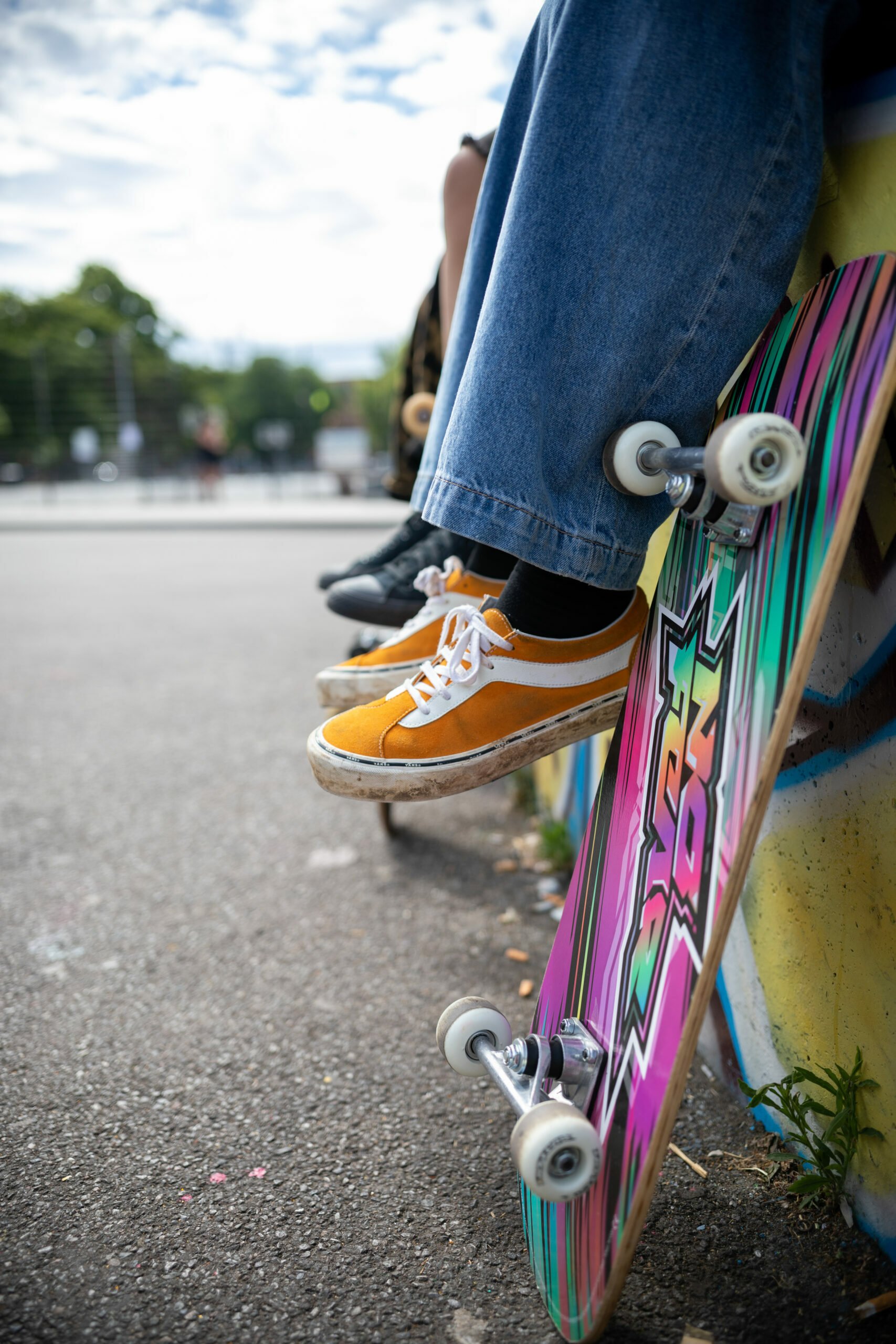 A close-up of a skateboard leaning against a wall with colorful graffiti. A person wearing orange and white sneakers and blue wide-legged jeans is sitting on the edge of the wall, with their feet dangling above the pavement. The skateboard has vibrant, multicolored stripes and visible white wheels. Other individuals and skateboards are slightly blurred in the background.