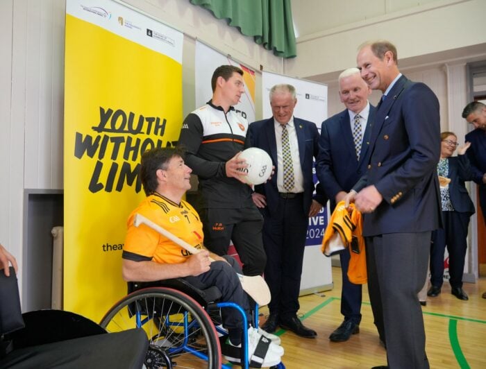 A group of five men are gathered in a gymnasium. One man in a wheelchair is holding a sports ball, while another man, standing beside him, is wearing a sports jersey. Two other men in suits are engaged in conversation with them, and a fifth man, dressed in a blazer, is smiling and interacting with the group. Behind them, there are banners promoting 