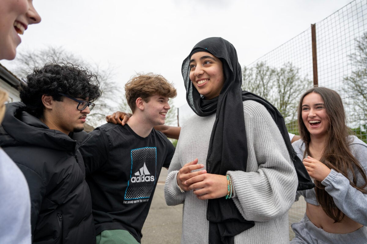 A group of teenagers stands closely together, smiling and laughing outdoors. One young woman wearing a grey sweater is in the center, smiling as she looks away. To her right, a girl with long brown hair, wearing a grey tracksuit, is laughing. On the left, a boy with curly black hair and glasses, dressed in a puffer jacket, stands next to another boy in a black Adidas t-shirt, both smiling and relaxed. The group appears to be enjoying each other's company in a casual, friendly moment.