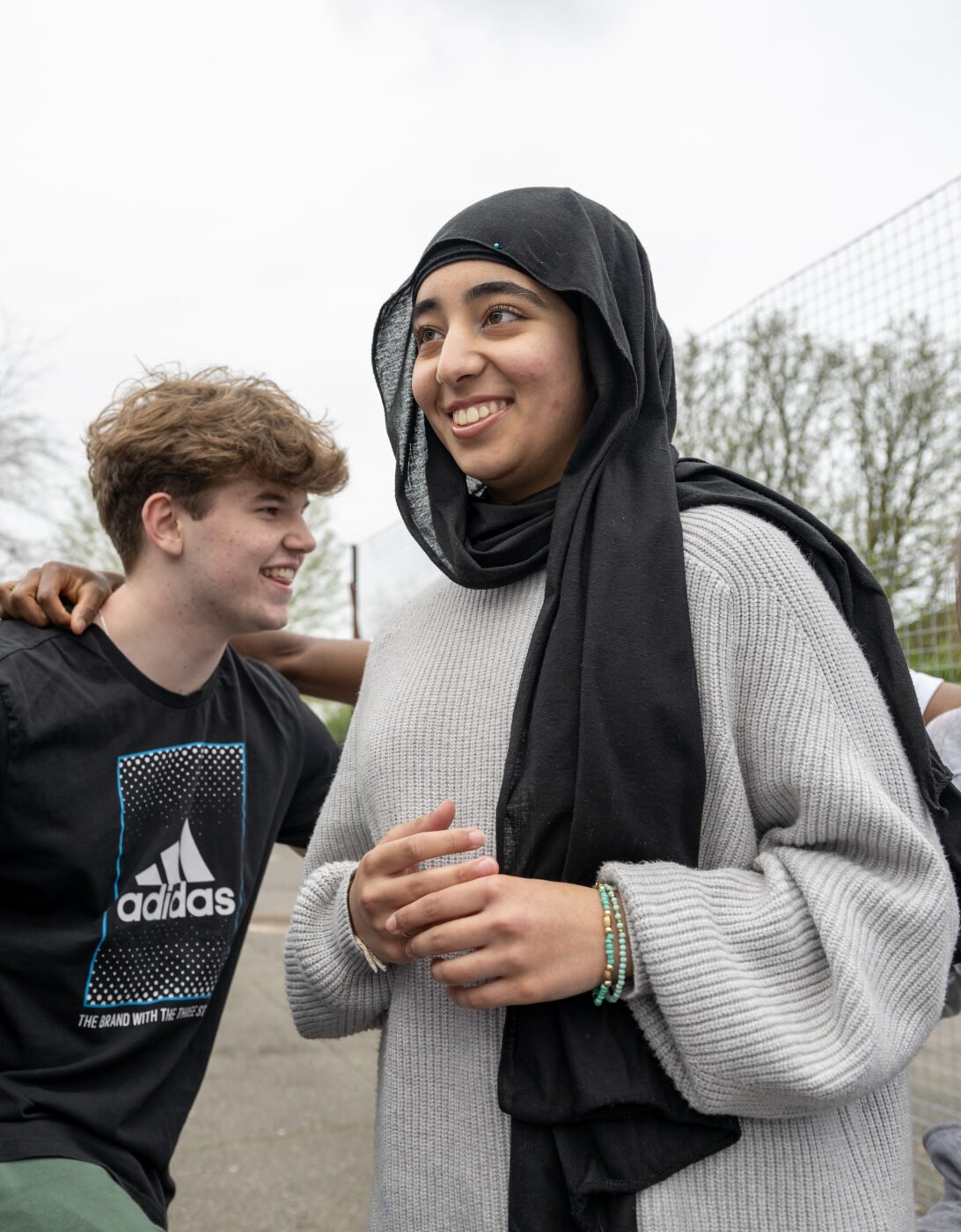 A group of teenagers stands closely together, smiling and laughing outdoors. One young woman wearing a grey sweater is in the center, smiling as she looks away. To her right, a girl with long brown hair, wearing a grey tracksuit, is laughing. On the left, a boy with curly black hair and glasses, dressed in a puffer jacket, stands next to another boy in a black Adidas t-shirt, both smiling and relaxed. The group appears to be enjoying each other's company in a casual, friendly moment.