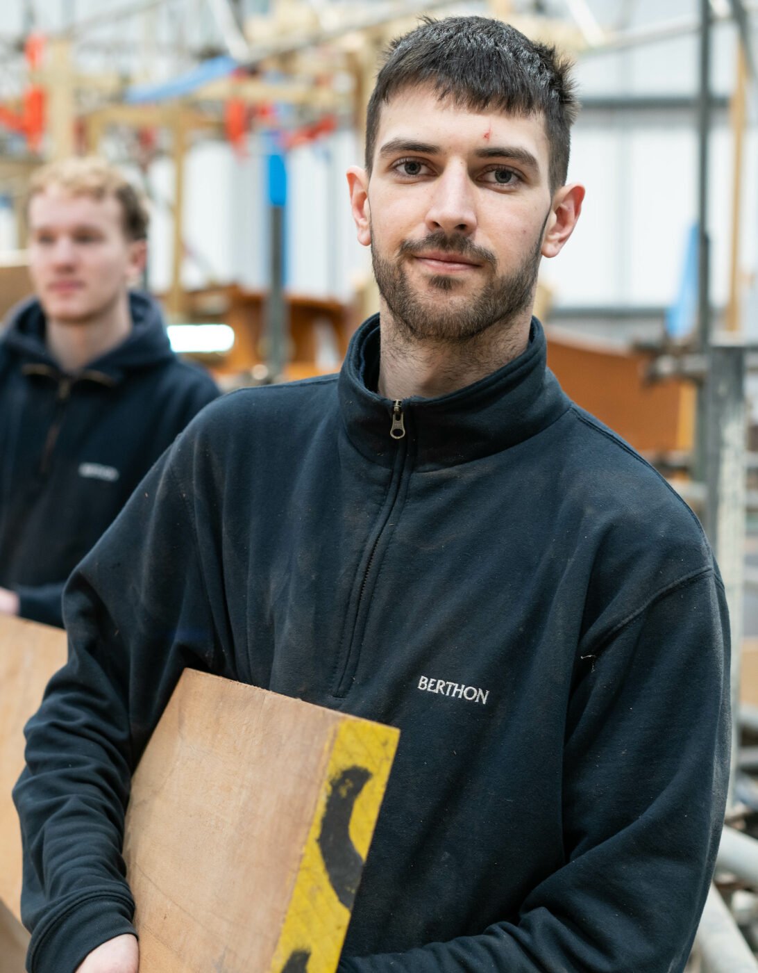 A group of young men working together in a workshop, focused on building a boat. The man closest to the camera has short dark hair, light facial hair, and is wearing a dark fleece jacket with the logo 