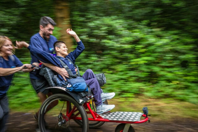 A boy with a joyful expression is being pushed in a specialized wheelchair along a wooded path. He is wearing a blue shirt and has his arms raised in excitement. Two adults, one in the foreground and one behind, are helping to push the wheelchair, both smiling and enjoying the moment. The background is a blur of green foliage, suggesting movement and speed.