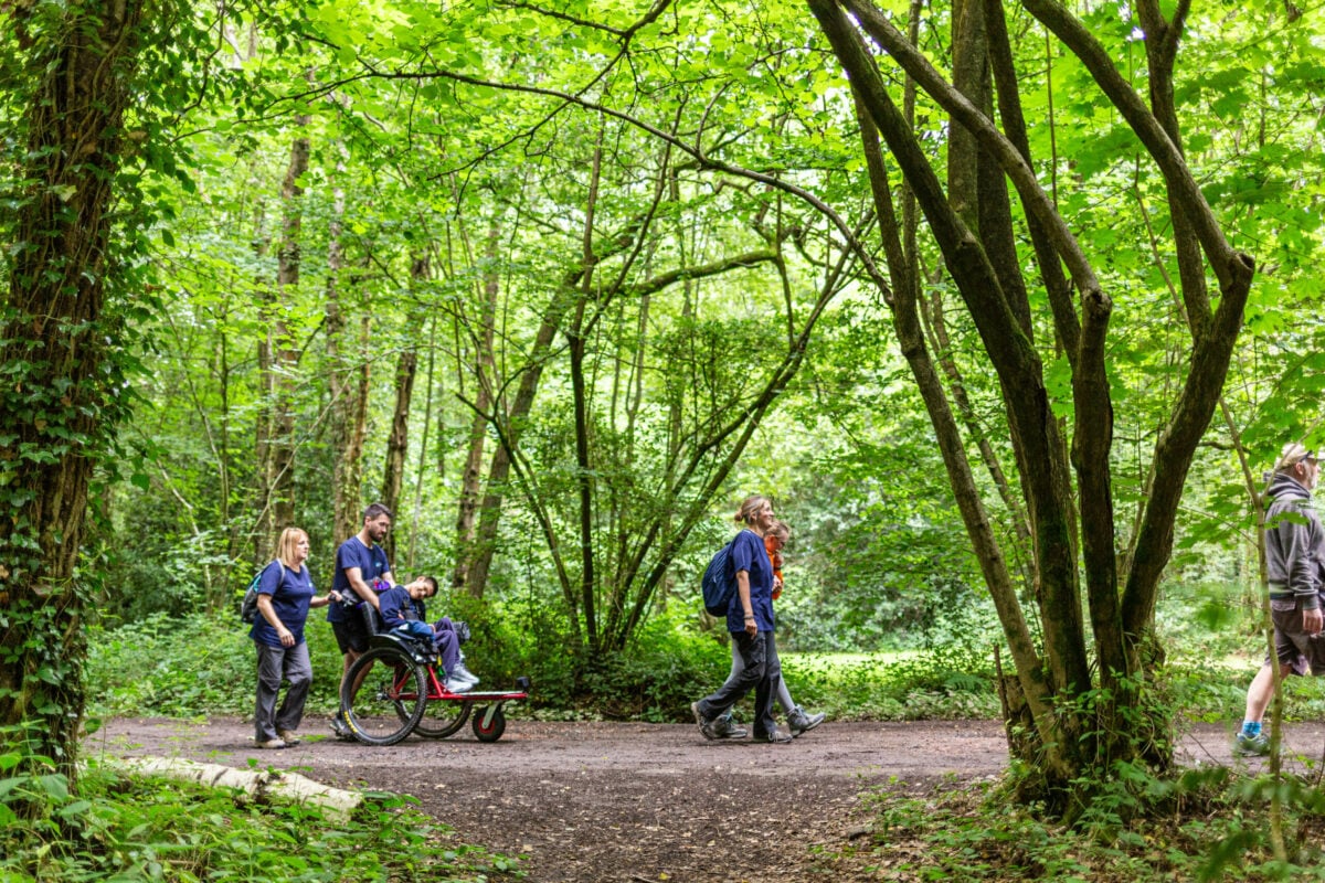 Two DofE Leaders assist Taylor in his wheelchair on a forest path. The woman is pushing the wheelchair while the man is smiling and engaging with Taylor. Lush greenery surrounds the path, and other individuals can be seen walking in the background.