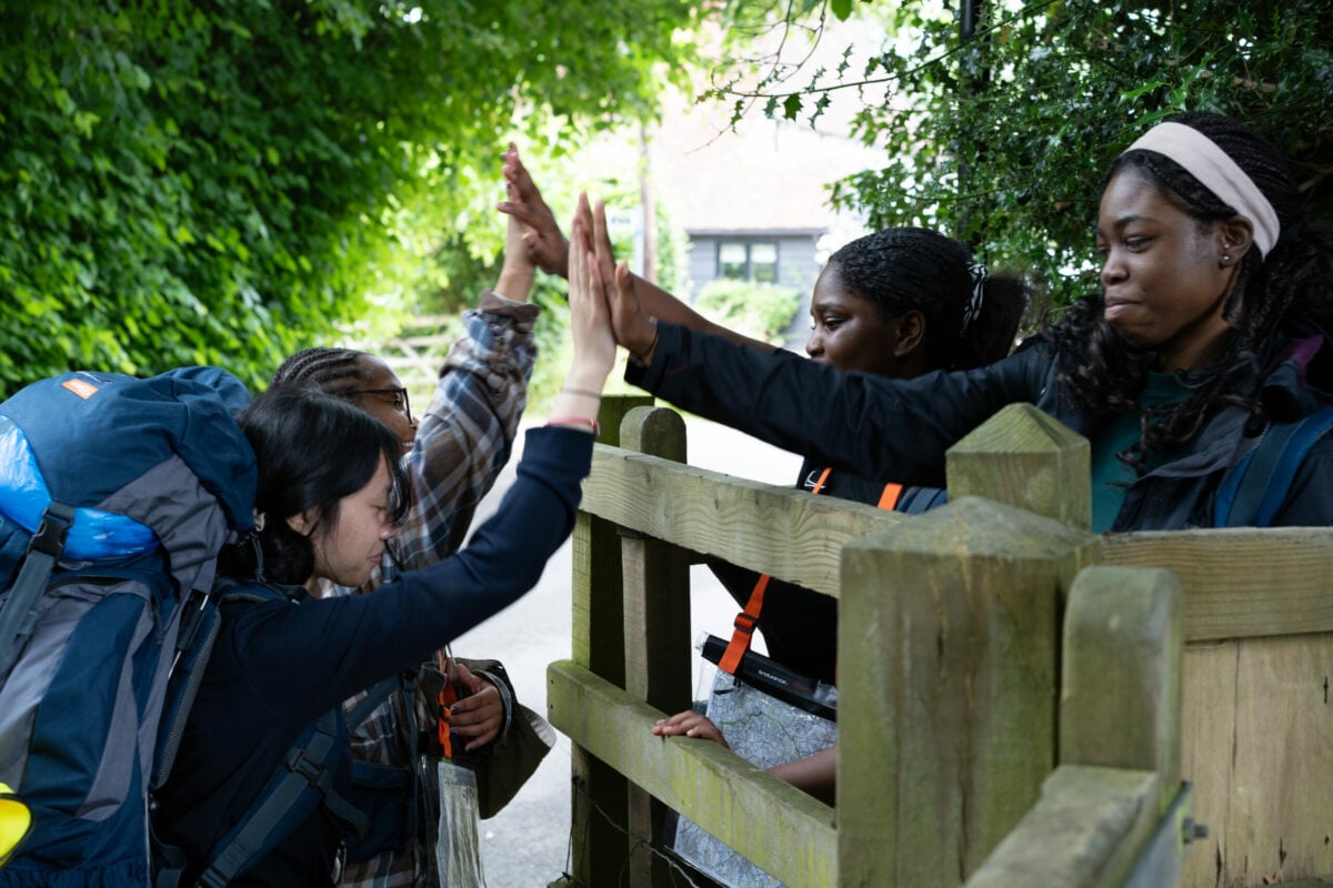 A group of young people on an outdoor expedition, wearing backpacks and outdoor clothing, are gathered by a wooden gate on a countryside path. Two individuals on one side of the gate are giving high-fives to two others on the opposite side. The setting is surrounded by lush green trees, creating a vibrant and lively atmosphere. Some participants are holding maps.