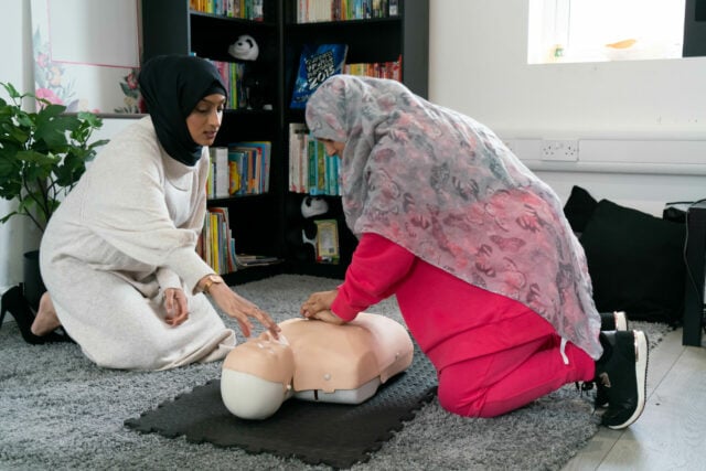 Two women are practicing CPR on a training mannequin in a cozy indoor setting. One woman is kneeling on the floor, performing chest compressions on the mannequin, while the other is beside her, offering guidance. The room has a calm atmosphere with bookshelves and soft furnishings in the background. Both women are focusing intently on the training.