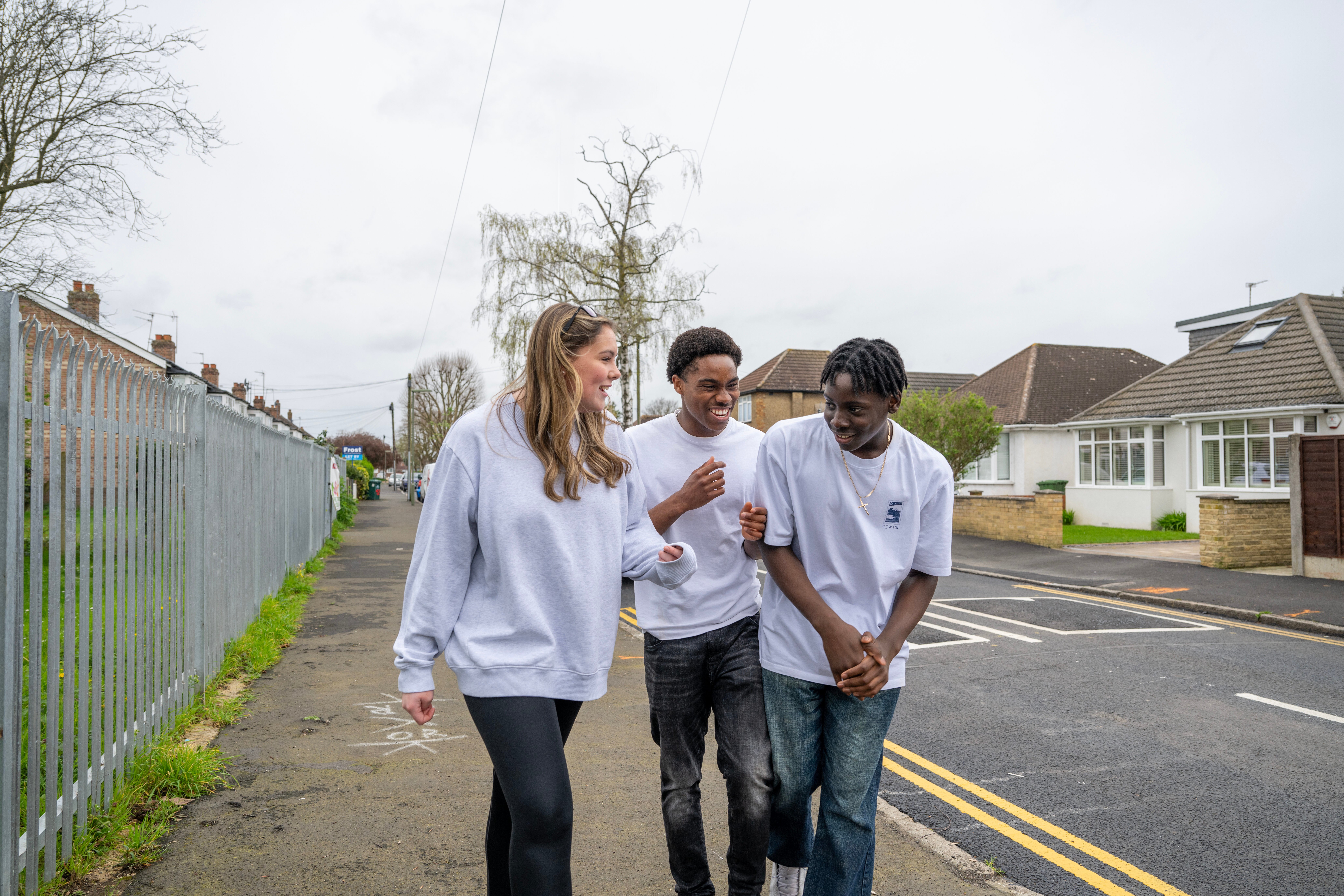 Three teenagers walking together and laughing on a suburban street. A girl in a grey sweatshirt is chatting with two boys, one wearing a white t-shirt and jeans, and the other in a white t-shirt and dark trousers. They appear to be enjoying each other's company on a cloudy day, with houses and a metal fence in the background.