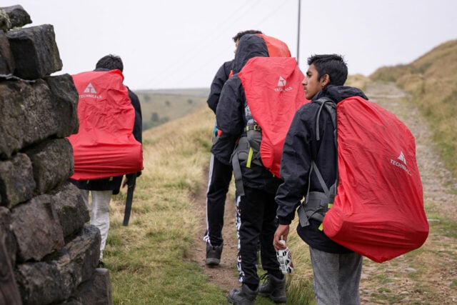 A group of young people walking along a rocky path in the countryside, carrying large red backpacks covered with rain covers labeled 'Technicals'. They are dressed in outdoor gear, including waterproof jackets, and one person holds a water bottle. The weather appears overcast, and the landscape is grassy and hilly.