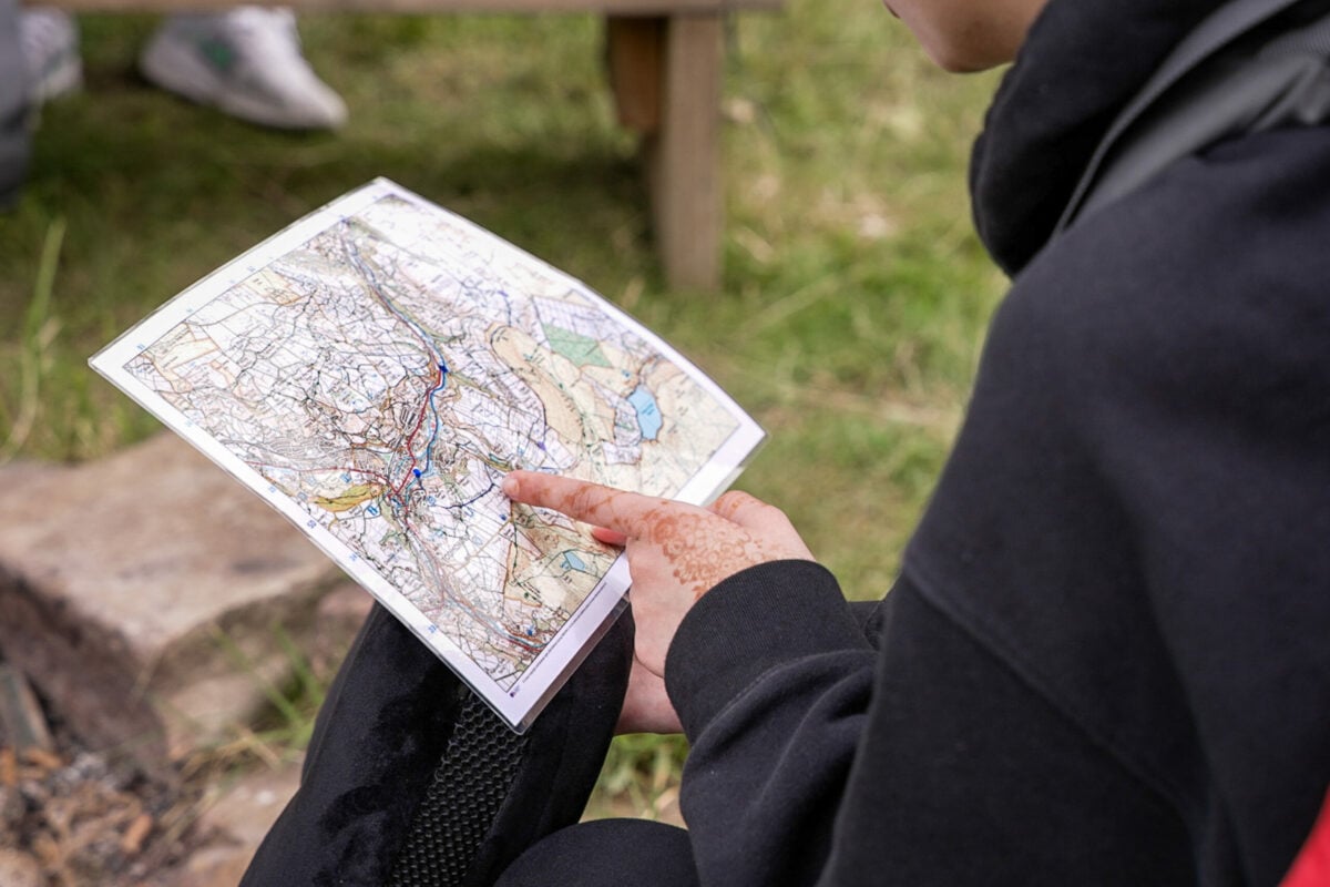 A person wearing a black jacket sits outdoors and holds a topographic map, pointing at a section of the map. The person's hand is adorned with henna. The setting appears to be a grassy area, and part of a wooden bench is visible in the background.