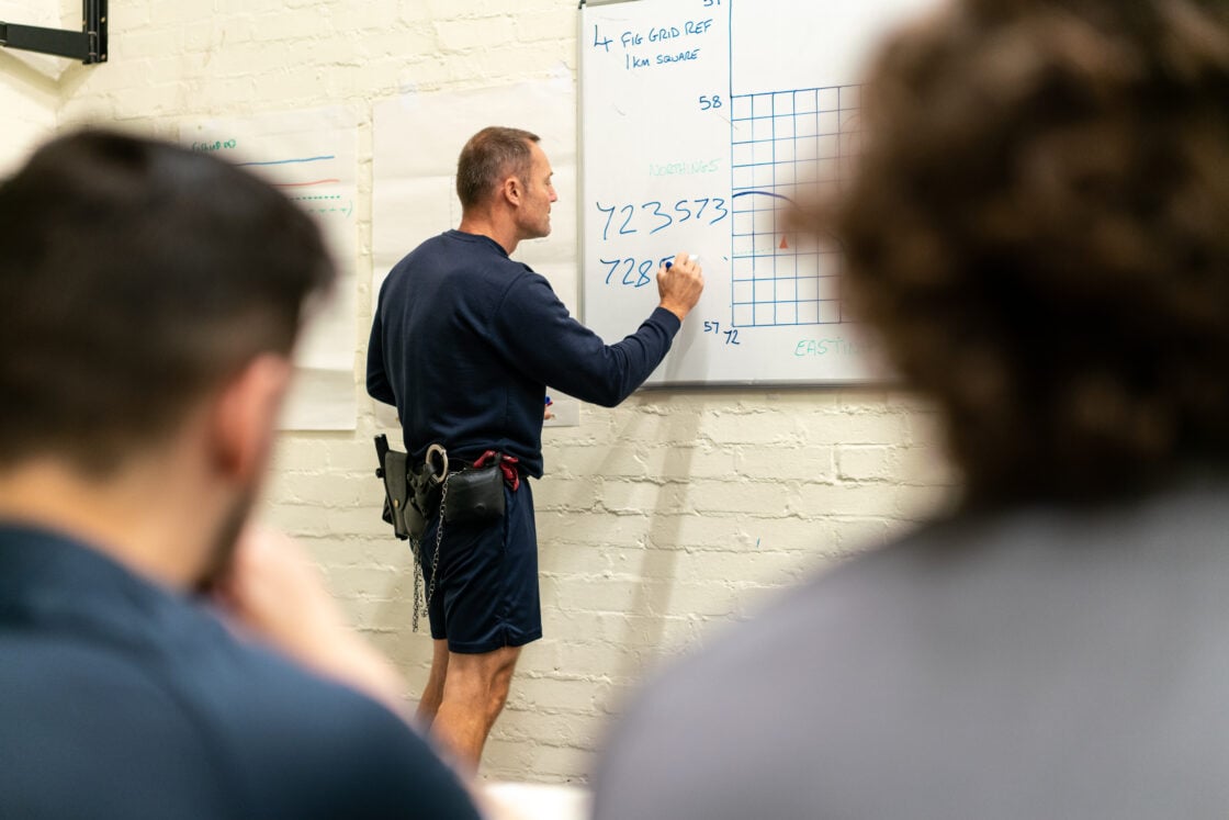 A man is standing at a whiteboard, writing coordinates and drawing on a grid. He is wearing a dark navy outfit with handcuffs, attached to his belt. Two other people are seated in the foreground, observing him, though their faces are out of focus. The man appears to be conducting a lesson or training session on map reading or navigation, as the whiteboard displays terms like 