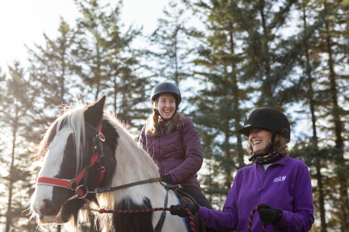 A young woman is riding a black-and-white horse, smiling broadly as she enjoys the experience. She is wearing a helmet and a purple jacket. Beside her, another woman, also wearing a helmet and a purple Riding for the Disabled Association (RDA) jacket, is leading the horse on a rope. Both are smiling and appear to be enjoying a pleasant outdoor ride, with tall pine trees in the background and sunlight filtering through the branches. The scene conveys a sense of joy and connection with nature.