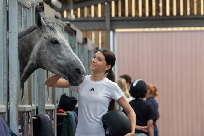 A young woman wearing a white Adidas t-shirt and holding a black riding helmet smiles while gently petting a gray horse in a stable. The horse leans its head toward her, and other people with helmets are visible in the background.