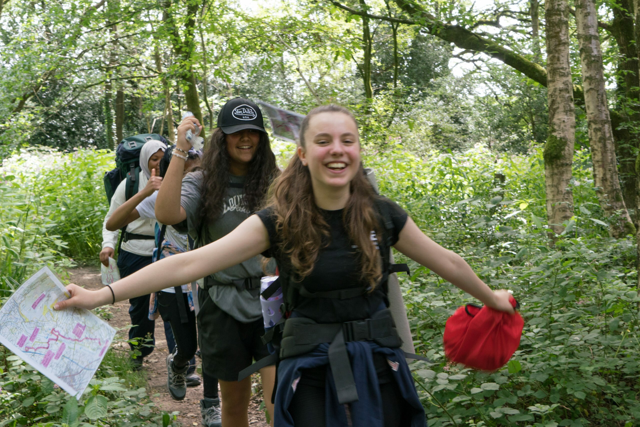 A group of young people hiking through a lush, green forest, all wearing backpacks. The person at the front, a smiling girl with long brown hair, has her arms spread wide in excitement, holding a red item in one hand and a map in the other. Behind her, two other participants follow, also smiling, one wearing a cap and the other a hoodie. Sunlight filters through the trees above.